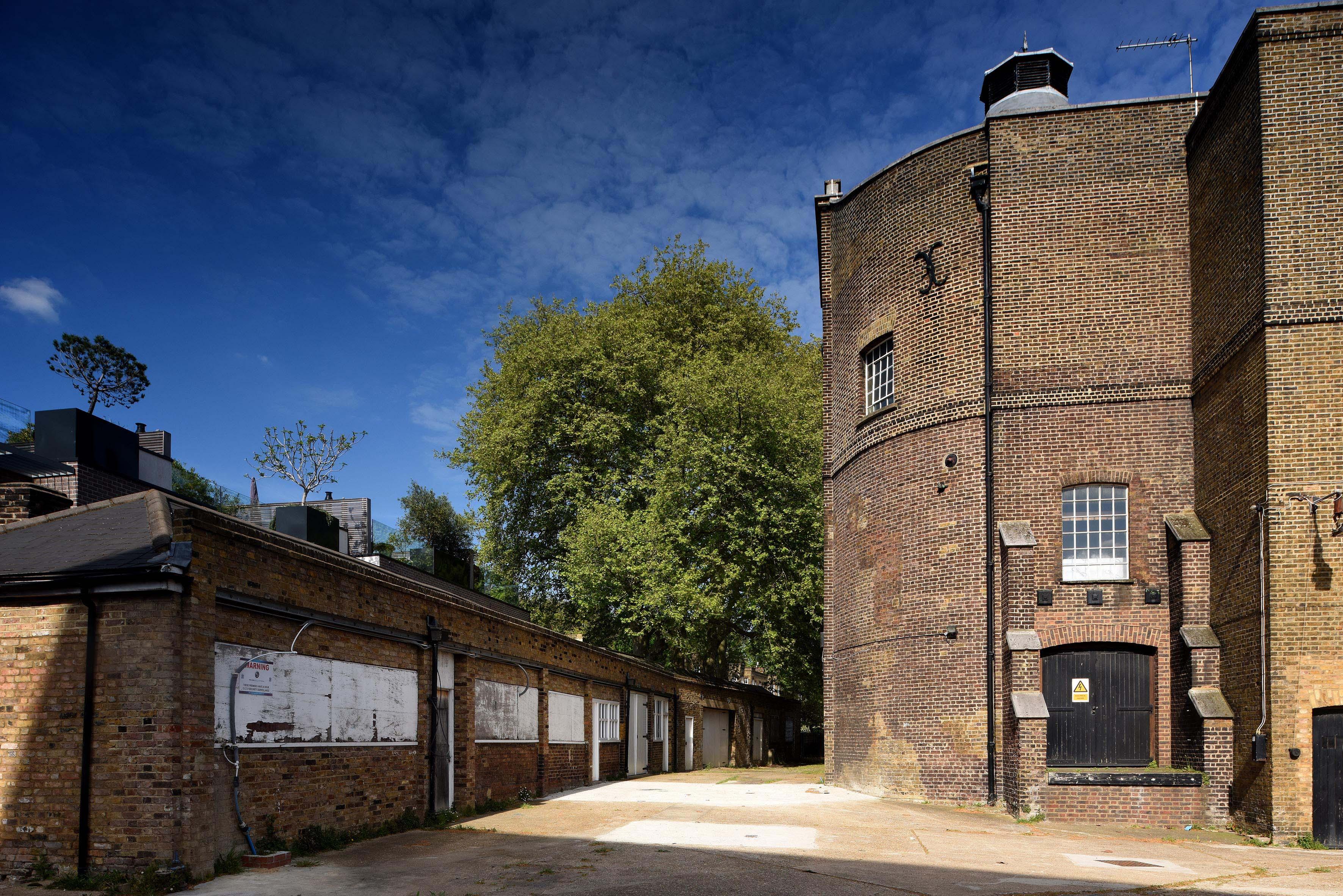 Photograph of a row of one story brick buildings on the left and a large derelict brick building on the right with a blue sky in the background.