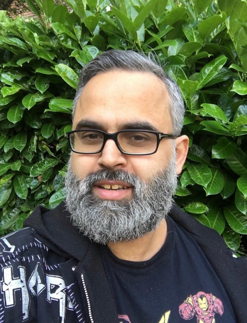A man with a grey beard and glasses and wearing a black t-shirt and hooded sweatshirt with graphics on, looks out towards the camera.
