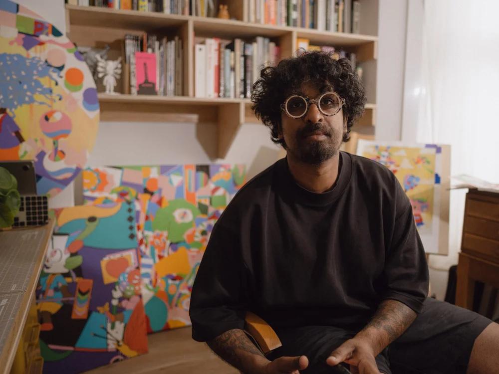 The artist MURUGIAH sits in his studio. He looks out towards the camera, behind him shelves of books and paintings.