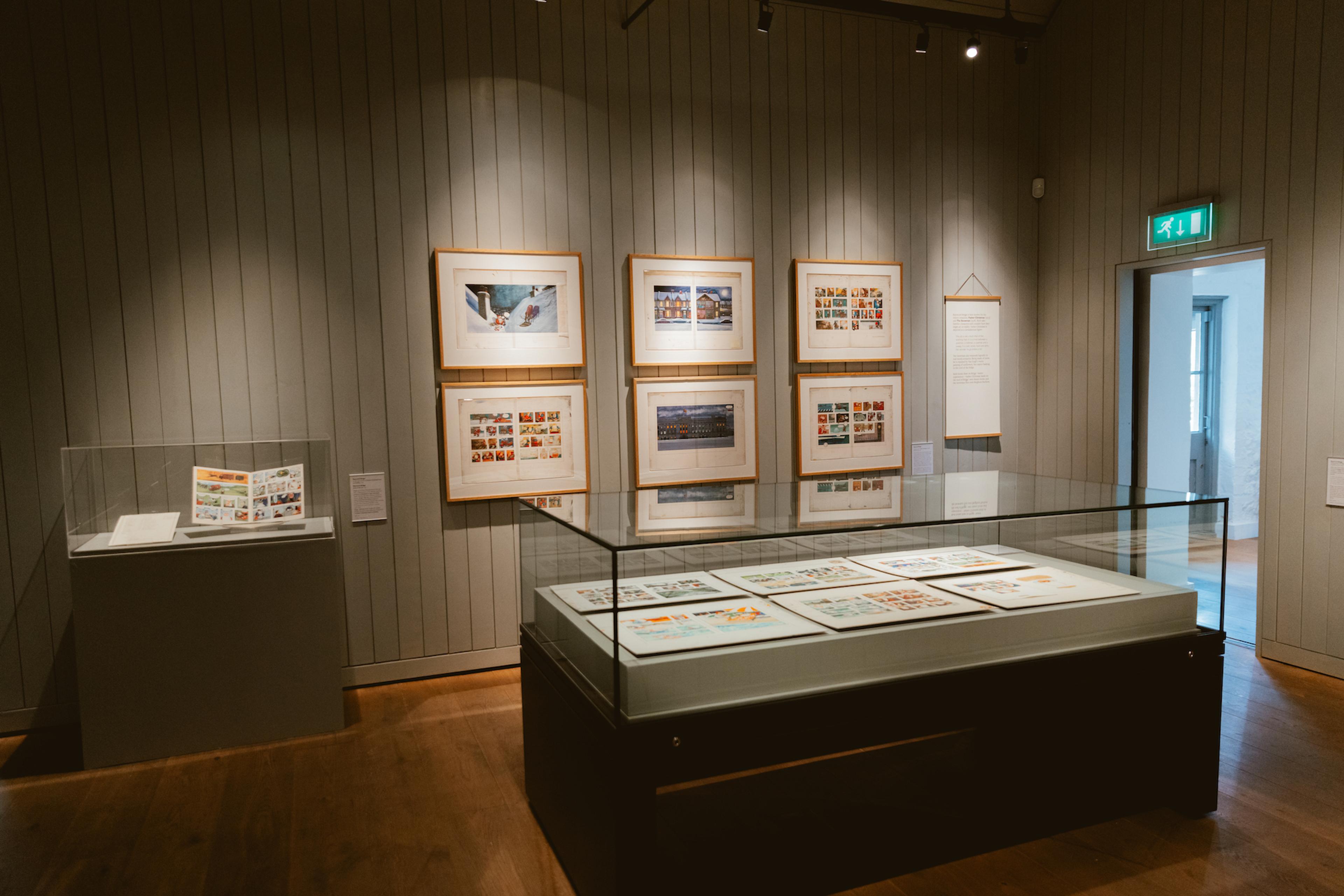 A photograph of a gallery with framed illustrations on the wall and a large glass display case in the centre of the room