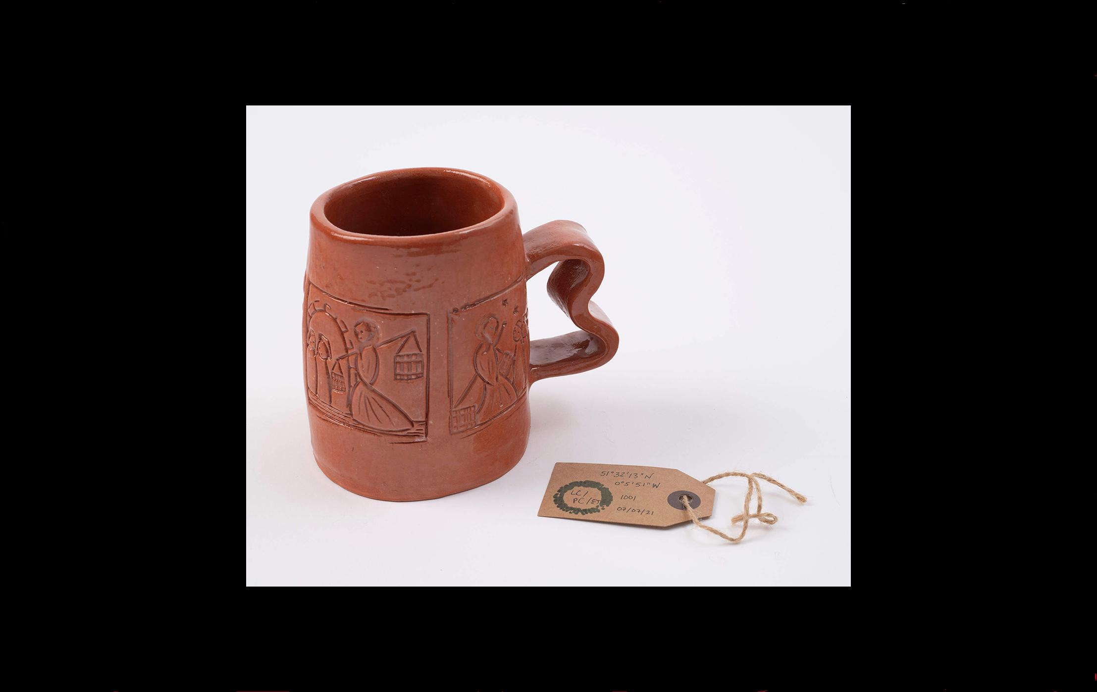 Photograph with a red clay mug with a brown museum label beside it. The mug is embossed using the wooden stamps displayed earlier in the sequence.