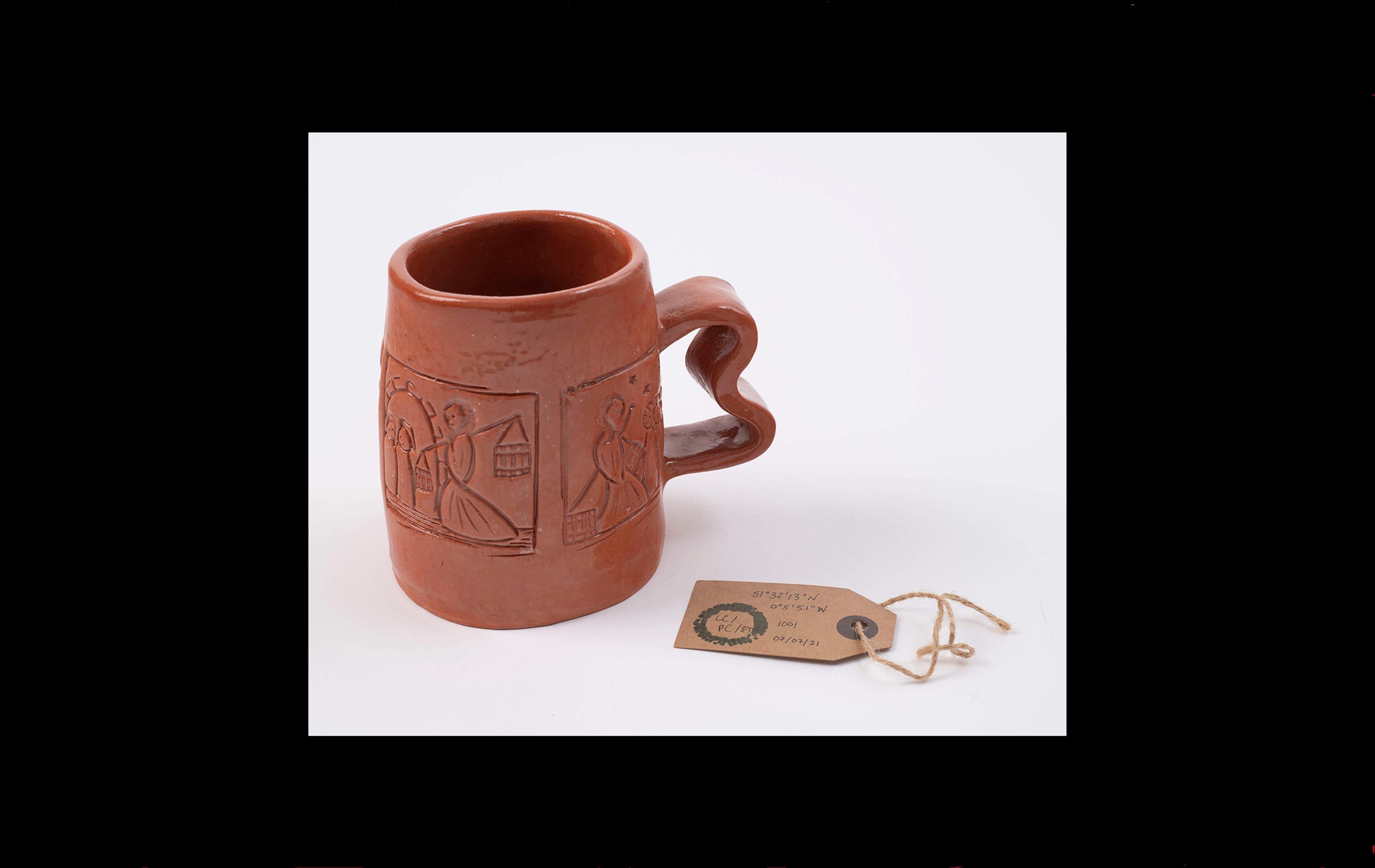 Photograph with a red clay mug with a brown museum label beside it. The mug is embossed using the wooden stamps displayed earlier in the sequence. 