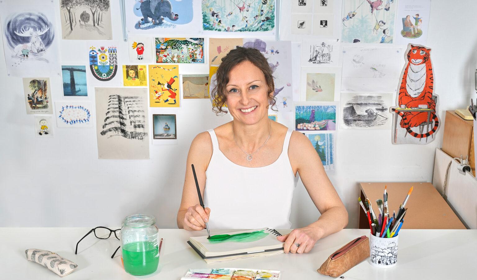 A woman sits in a studio with images pasted on the wall behind. She holds a paintbrush and is painting using green watercolour.