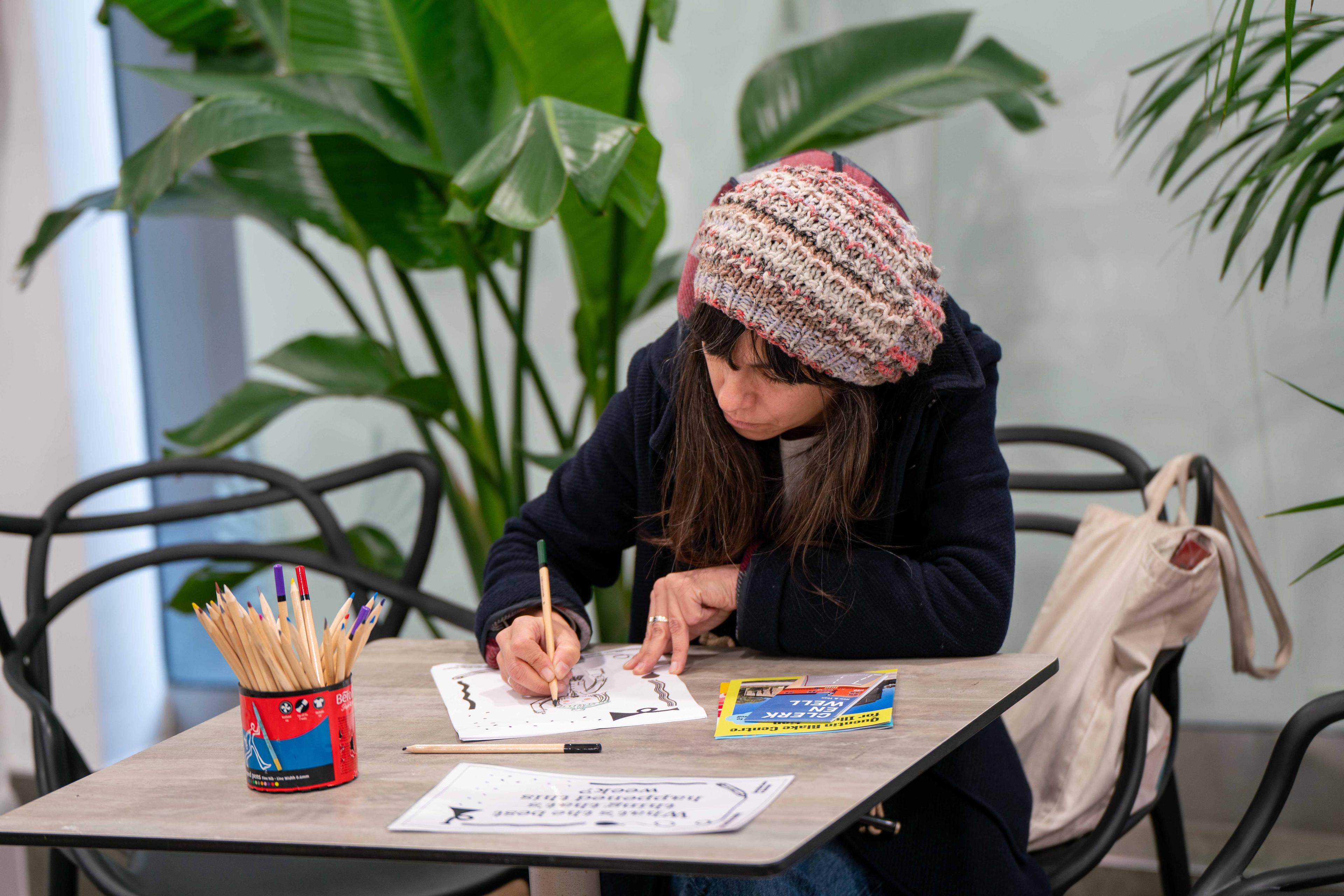 Photograph of a person at a desk drawing with pencils on a piece of paper on a table.