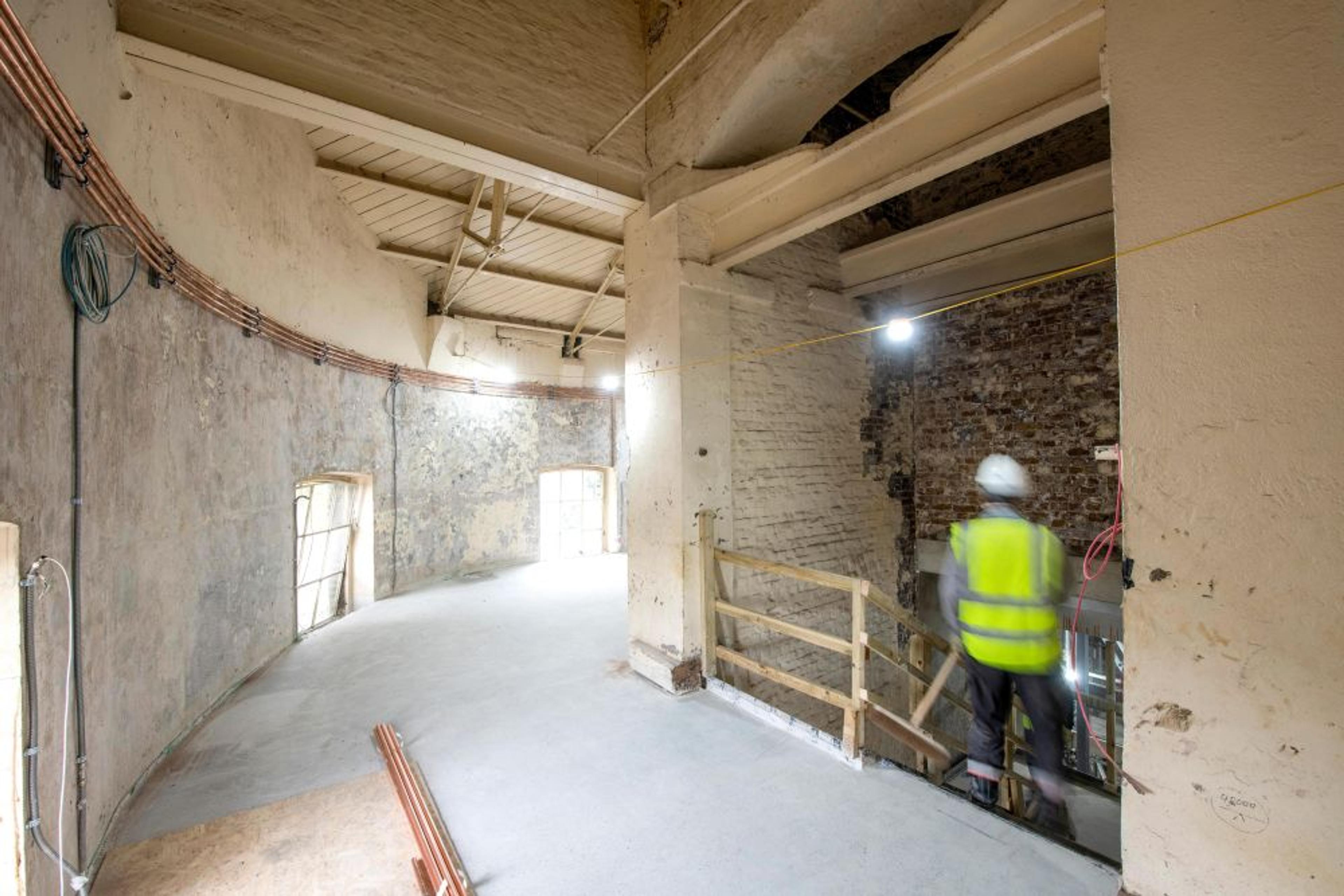 Interior of a curved industrial space under renovation. There are exposed brick walls, high ceilings, and a worker in a hard hat and reflective vest.