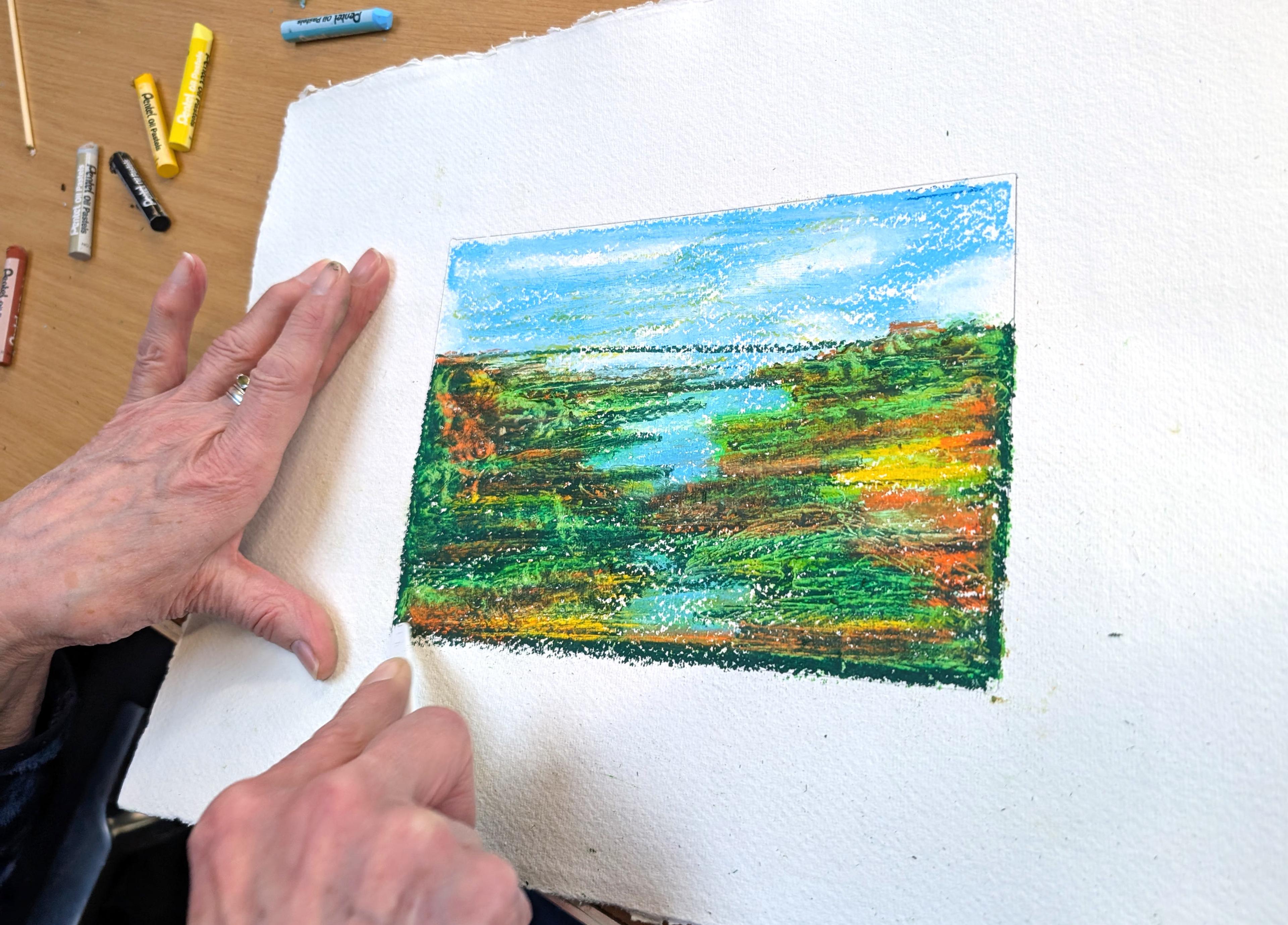 A piece of paper with an oil pastel drawing of a landscape. An adult's hand is holding a plastic tool which they are using to scratch details into the layers of oil pastel.