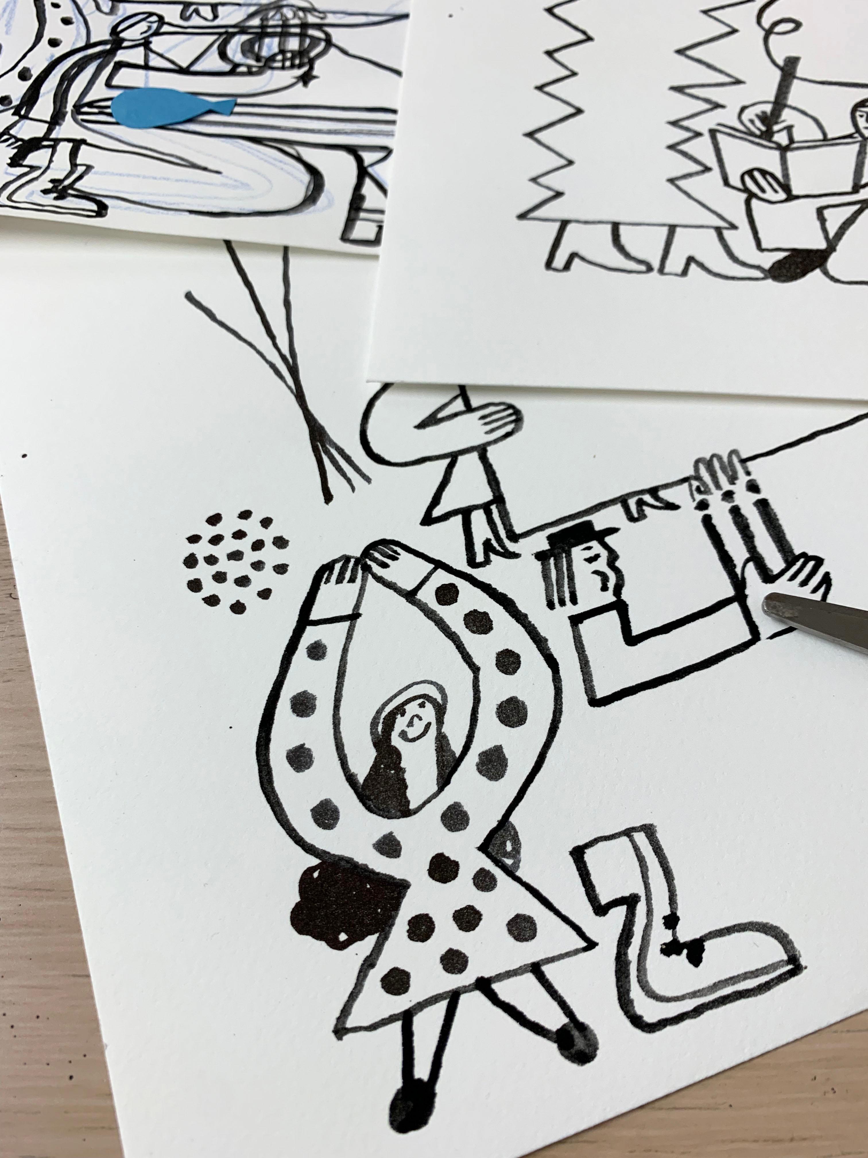 Black and white illustrations on a table surrounded by paint brushes, paper cut outs and scissors.