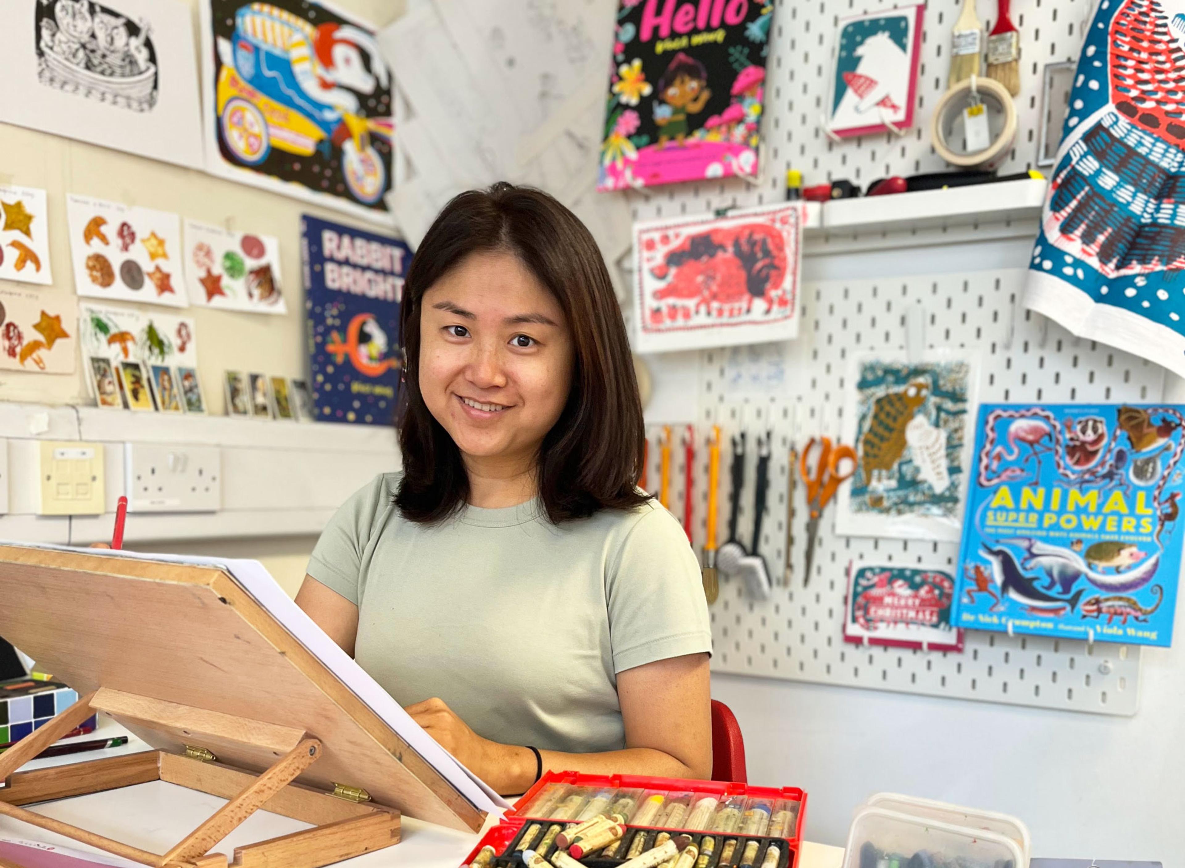 A person sits at a desk, drawing. Behind them are illustrations on display. 