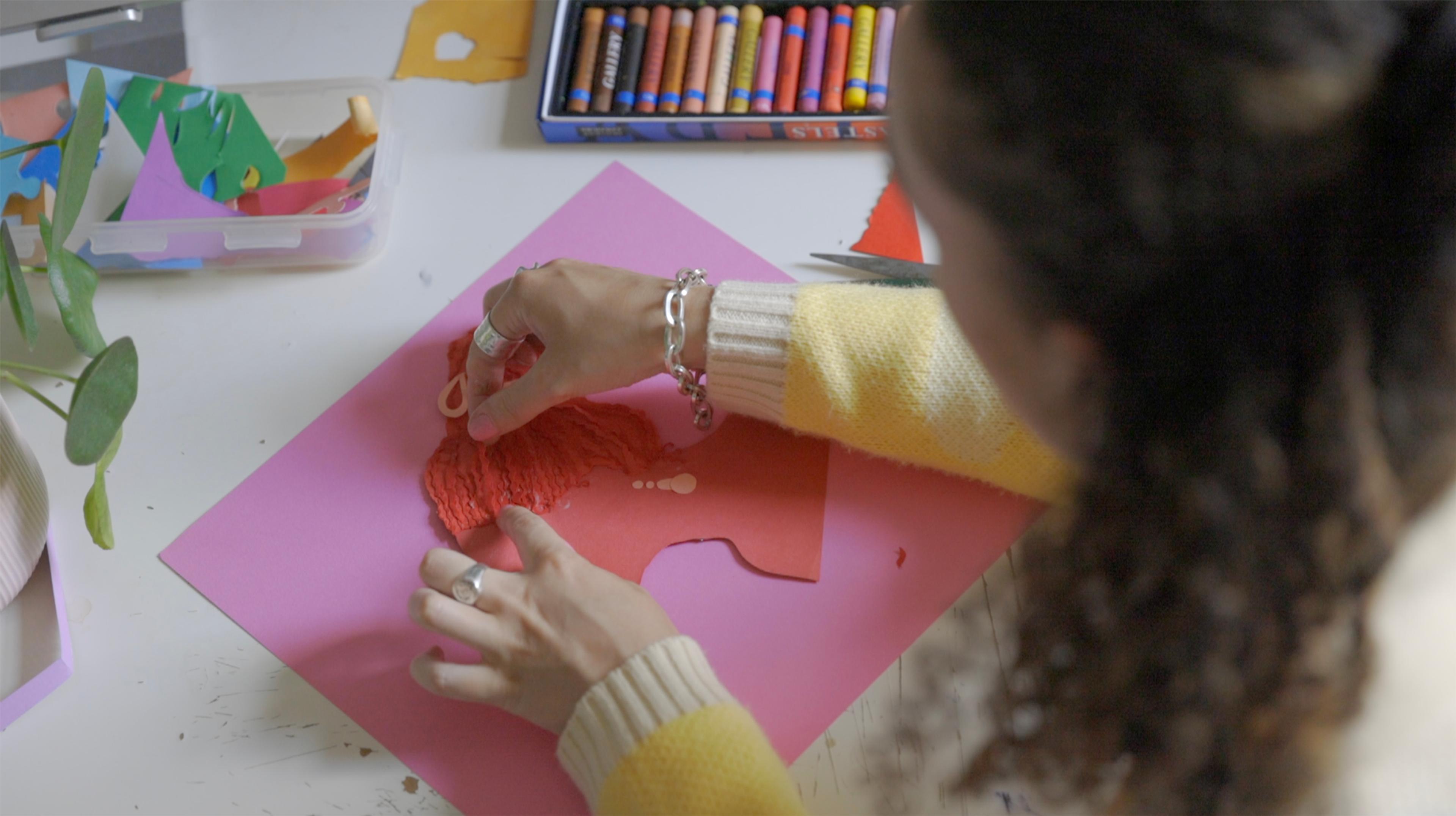 Photograph of a person collaging a paper portrait at a desk, there are crayons, a tub of cut out bits of colourful paper and a plant on top of it.