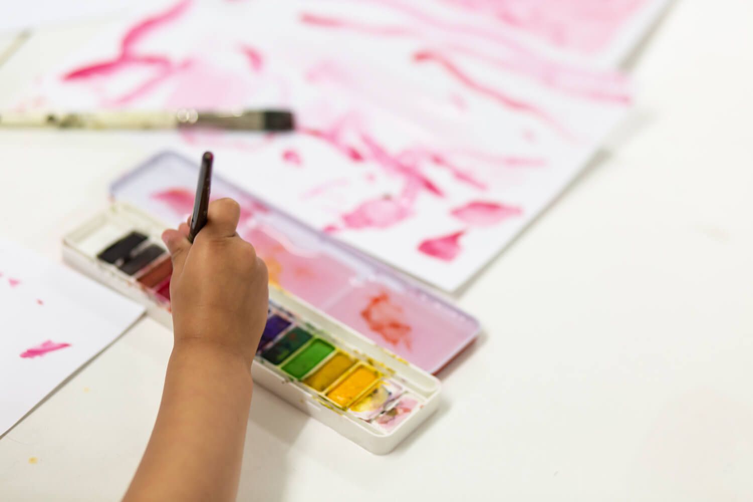 A photo of a child's hand holding a paintbrush that is being dipped into a watercolour paint palette with paint all the colours of the rainbow.