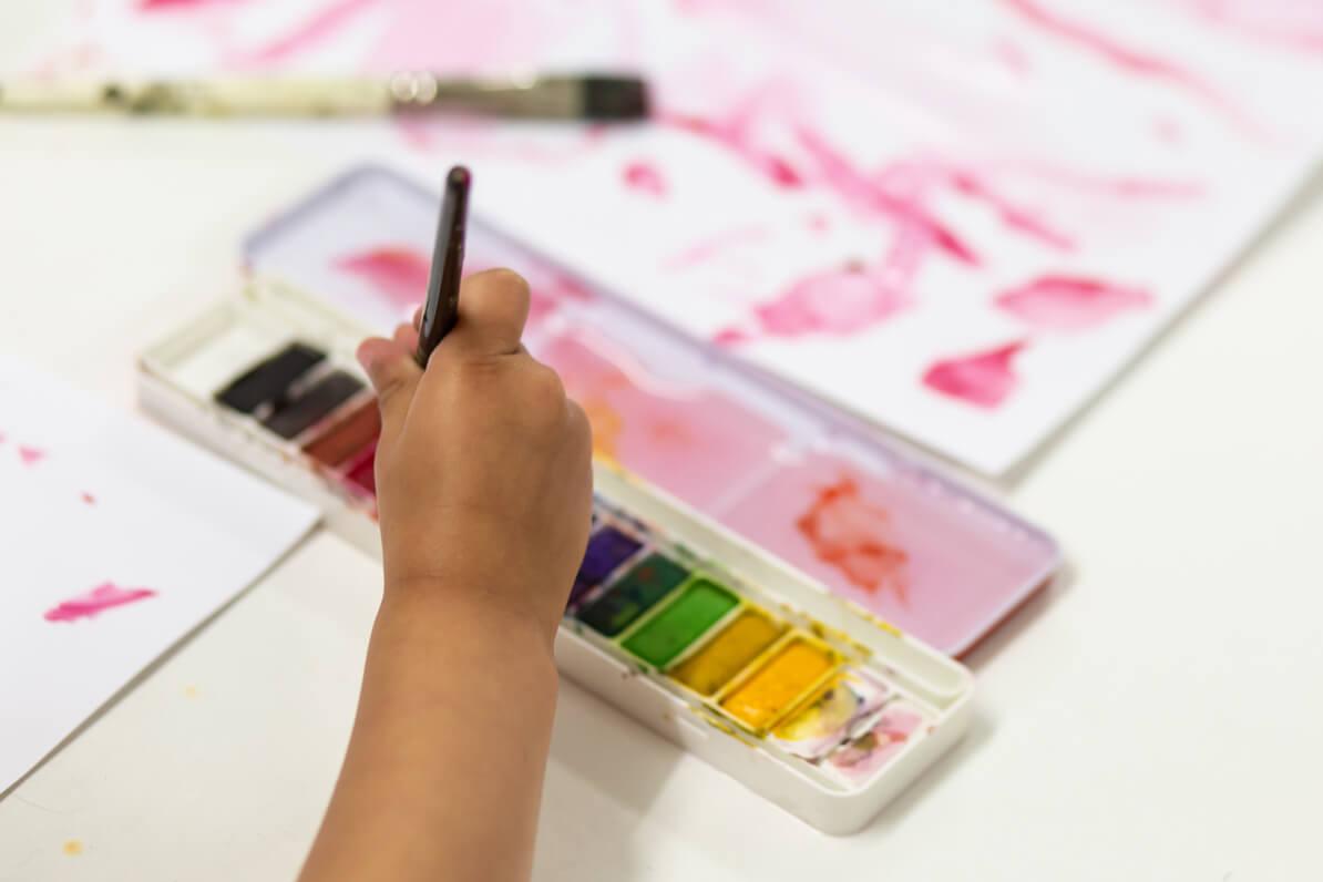 A photo of a child's hand holding a paintbrush that is being dipped into a watercolour paint palette with paint all the colours of the rainbow.