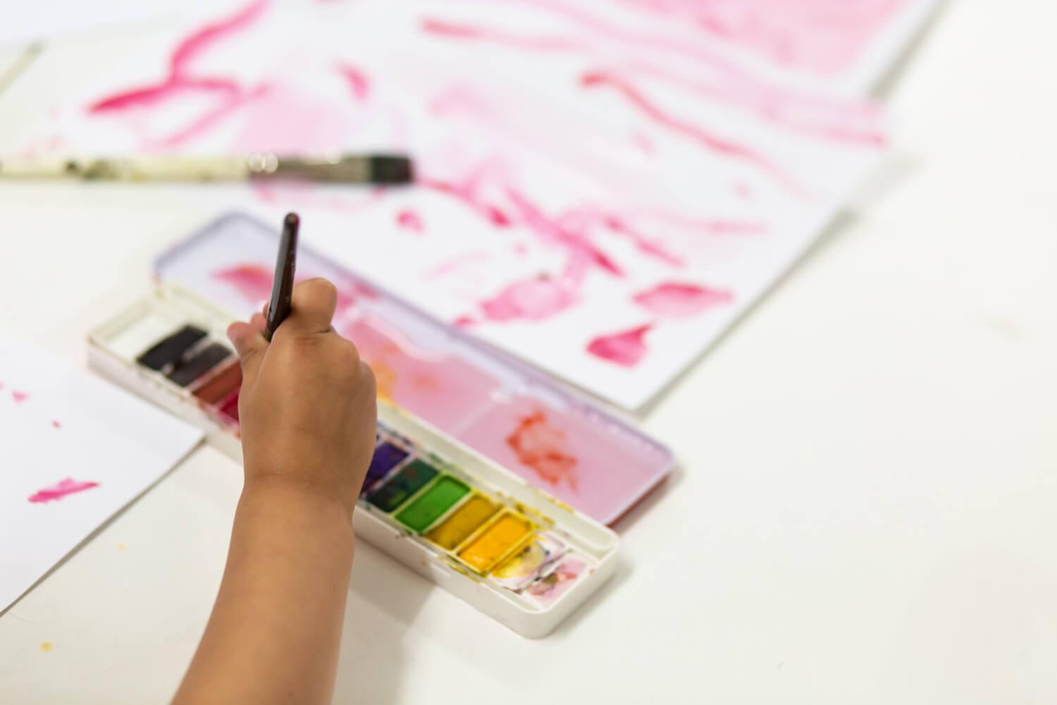 A photo of a child's hand holding a paintbrush that is being dipped into a watercolour paint palette with paint all the colours of the rainbow.