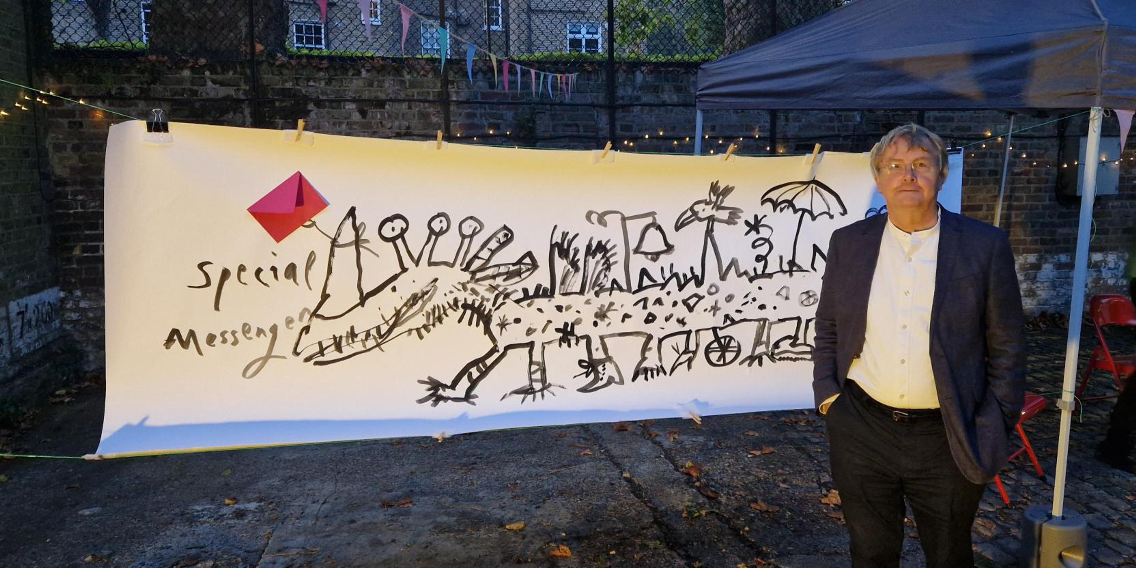 A photograph of the architect Tim Ronalds standing in front of a large banner with an illustration of a fantastical dragon-like creature holding a red envelope with the words "Special Messenger" 