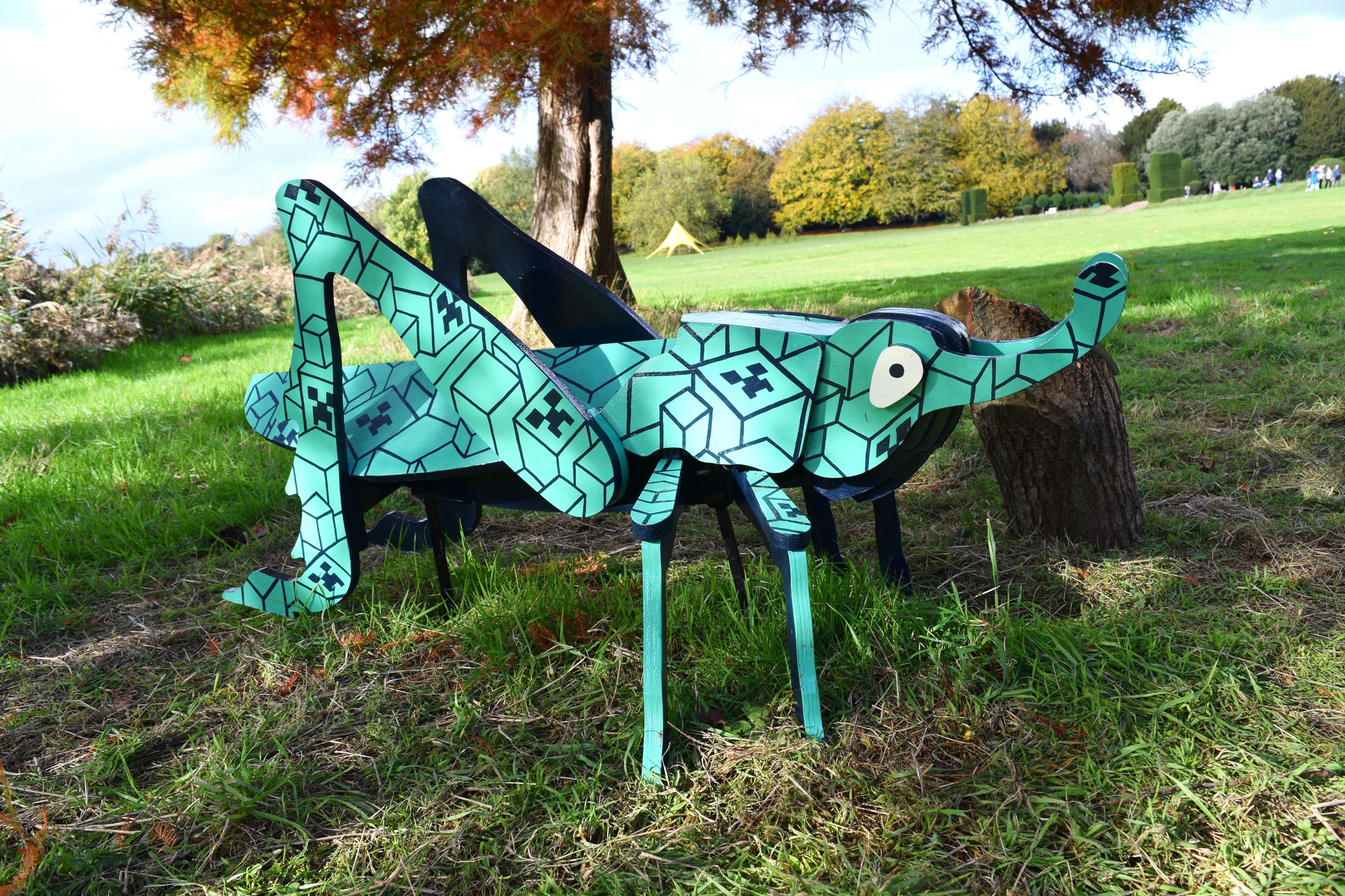 Photograph of a large grasshopper sculpture in a park, the grasshopper has been painted in green with a black pattern.