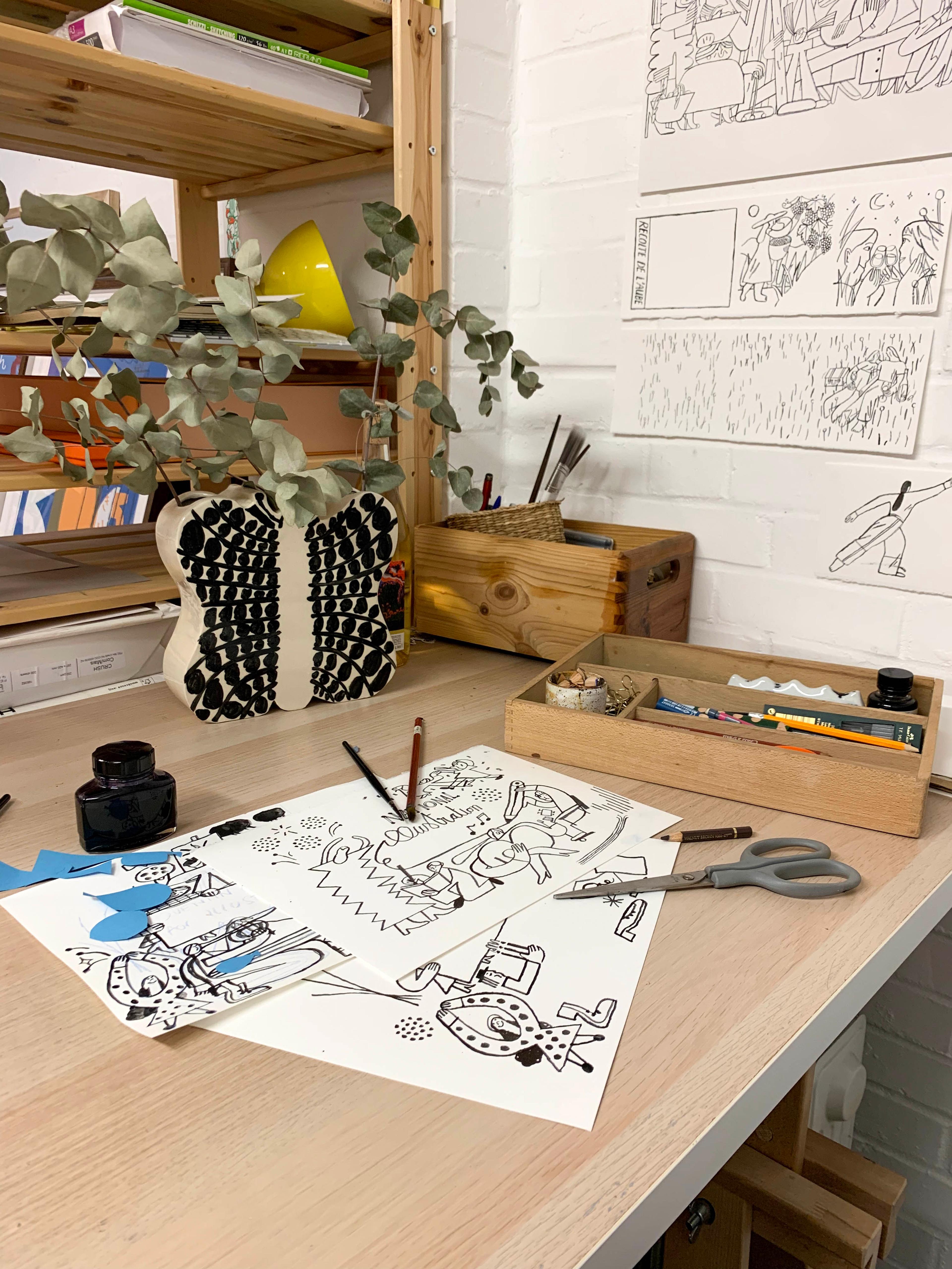 A photograph of a studio desk with illustrations on the table, with scissors, pens and ink.