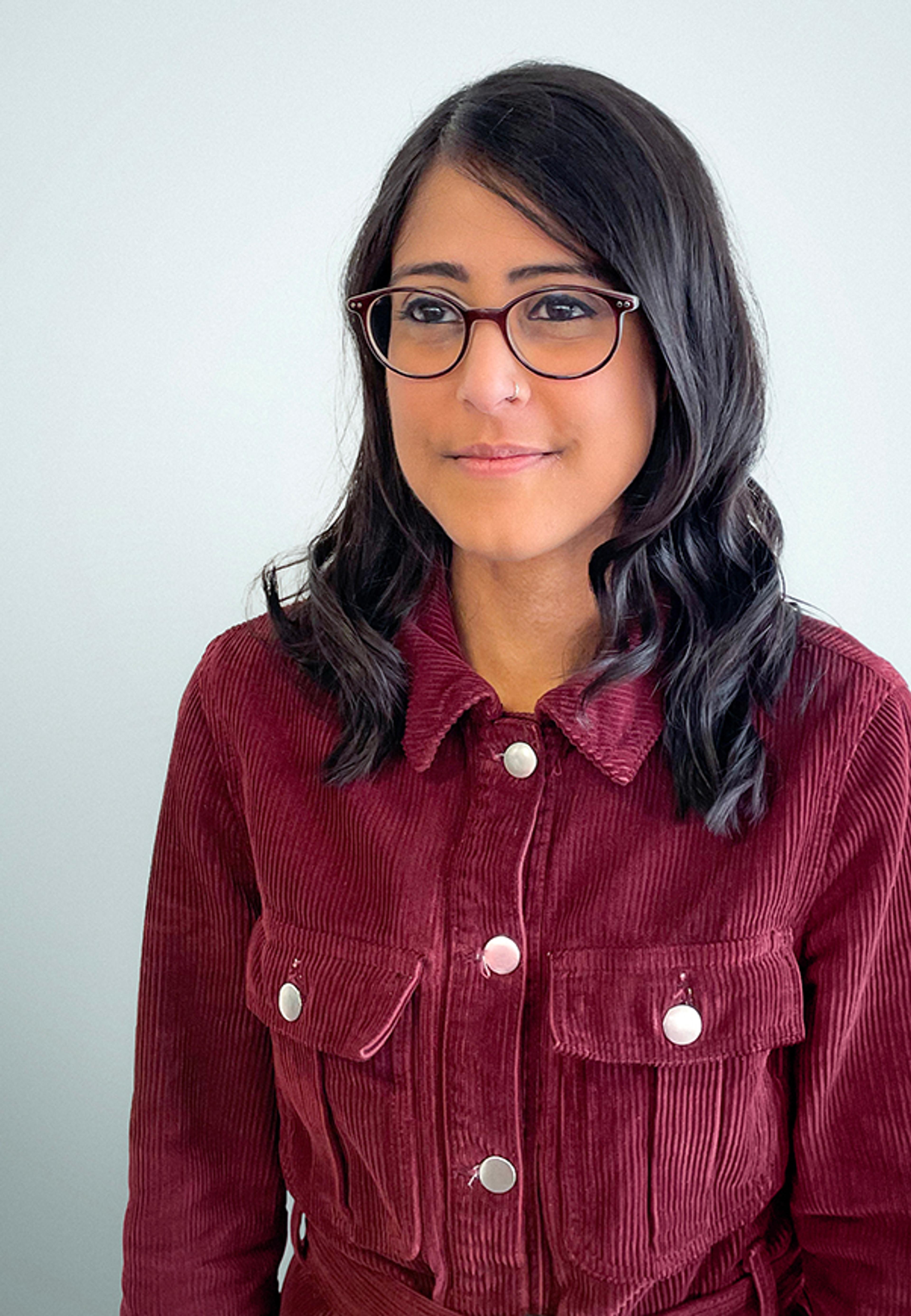 A woman with glasses and dark hair wearing a maroon corduroy jacket, smiles softly against a plain light background.
