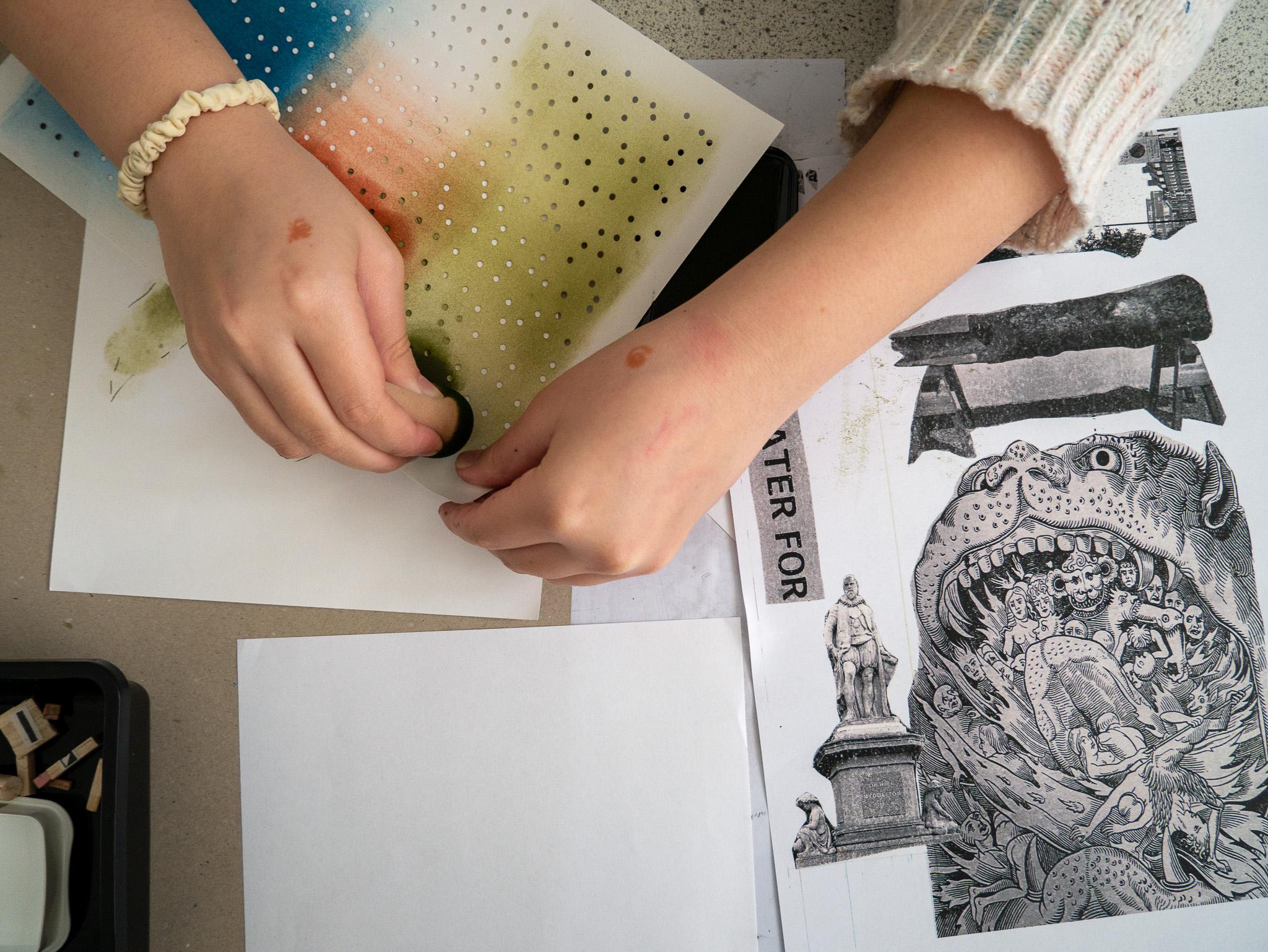 Two hands poke holes into a colourfully painted piece of paper besides a collage involving a statue and a beast with an open mouth and screaming figures inside.