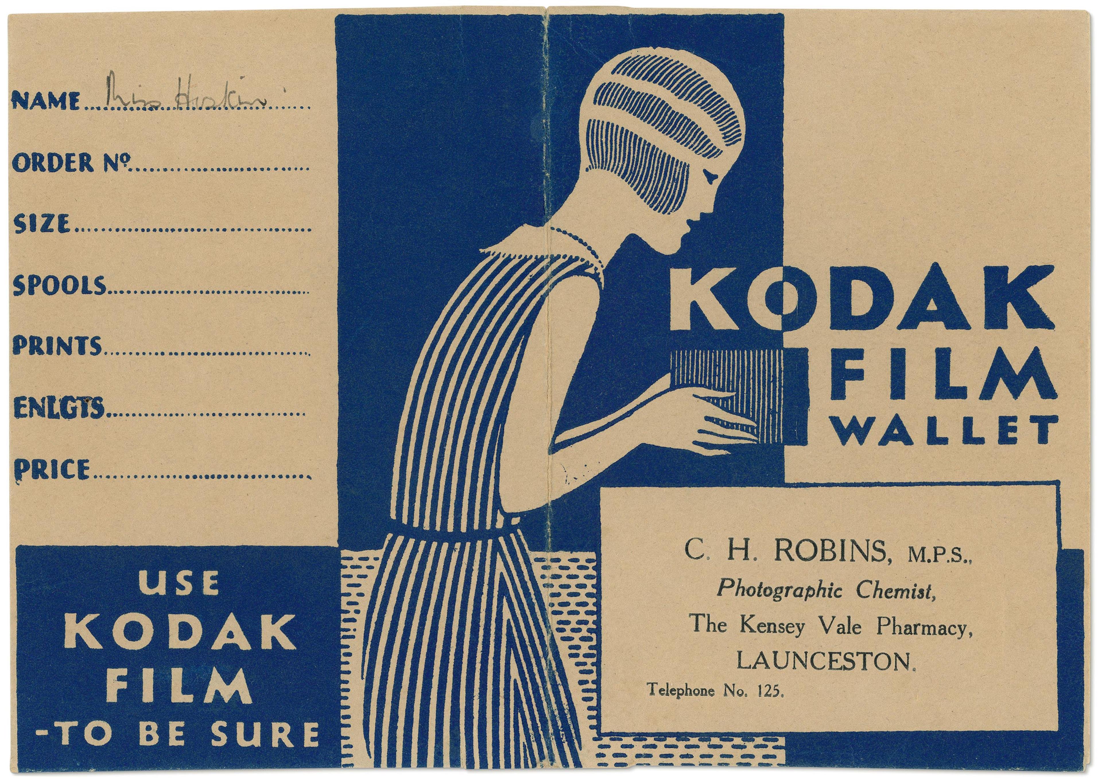 Photo of a paper envelope featuring a blue and white illustration of a person holding an envelope with the text 'Kodak film wallet' 'use Kodak film - to be sure', along with a space to write details such as name, order number, size etc. along with the address of 'C. H. Robins M.P.S Photographic Chemist'.