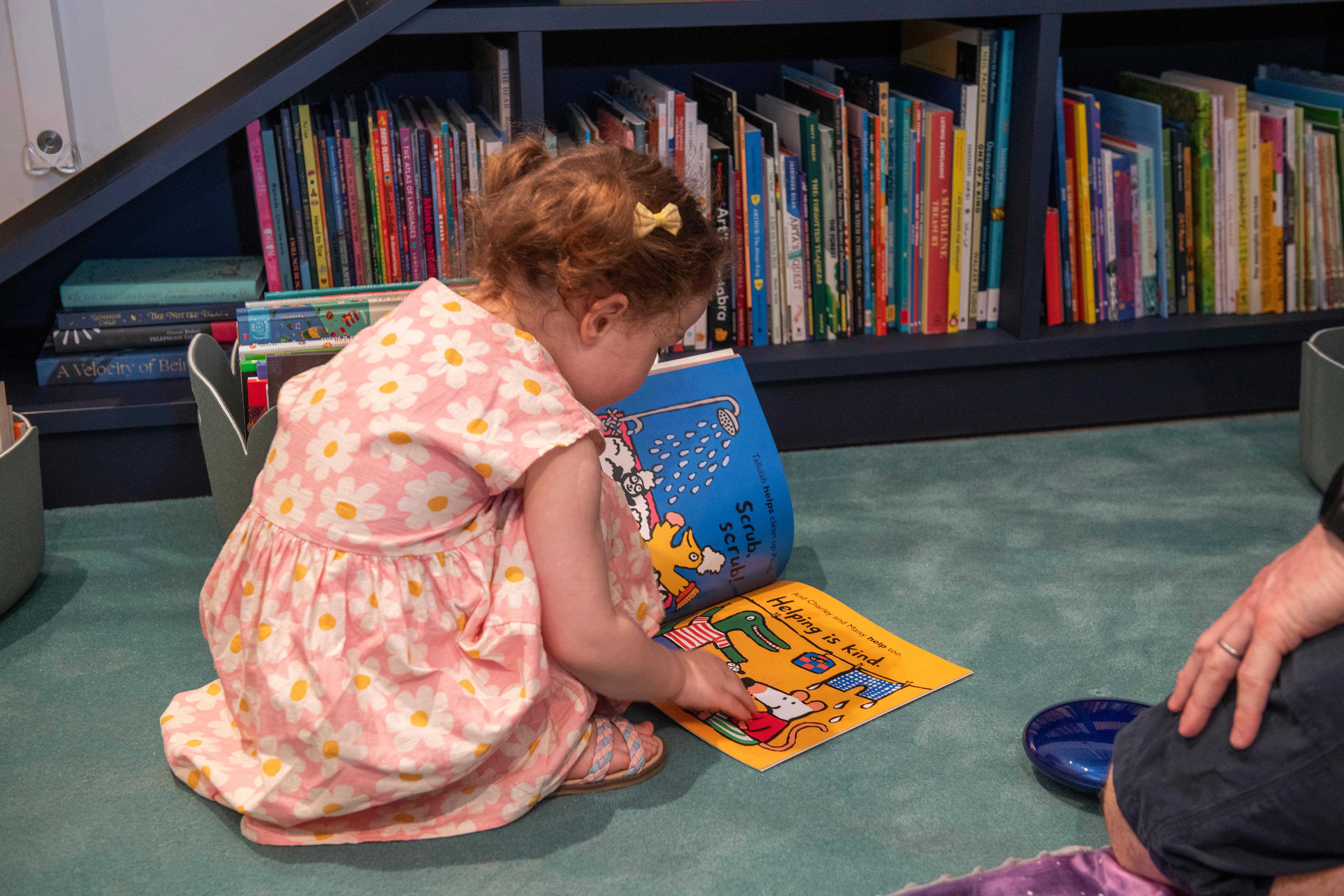 A photo of toddler in a pink and white flower print dress reading from a colourful picture book in a carpeted reading nook