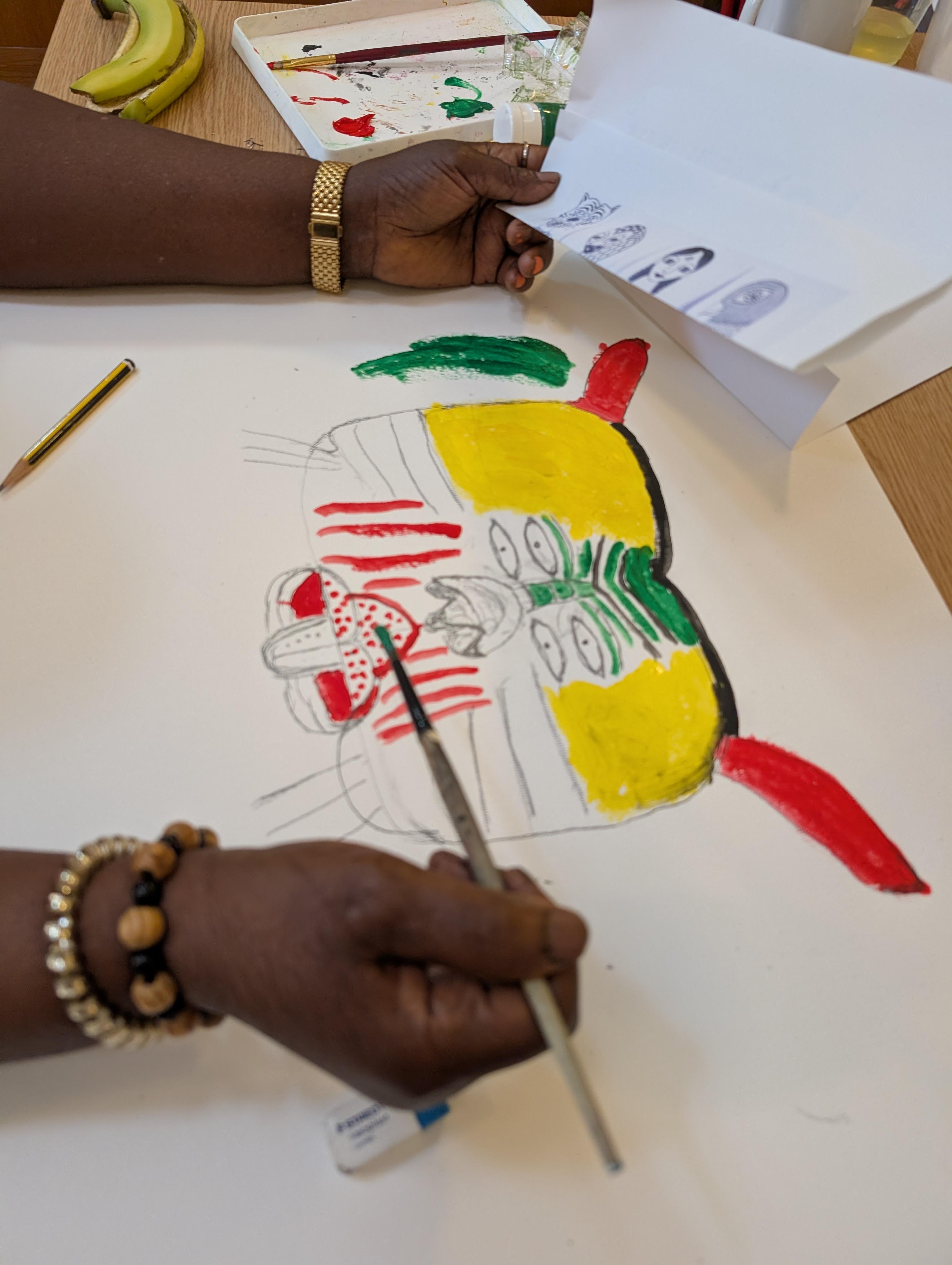A person's hand painting a mask-like face with four eyes and bright colours.