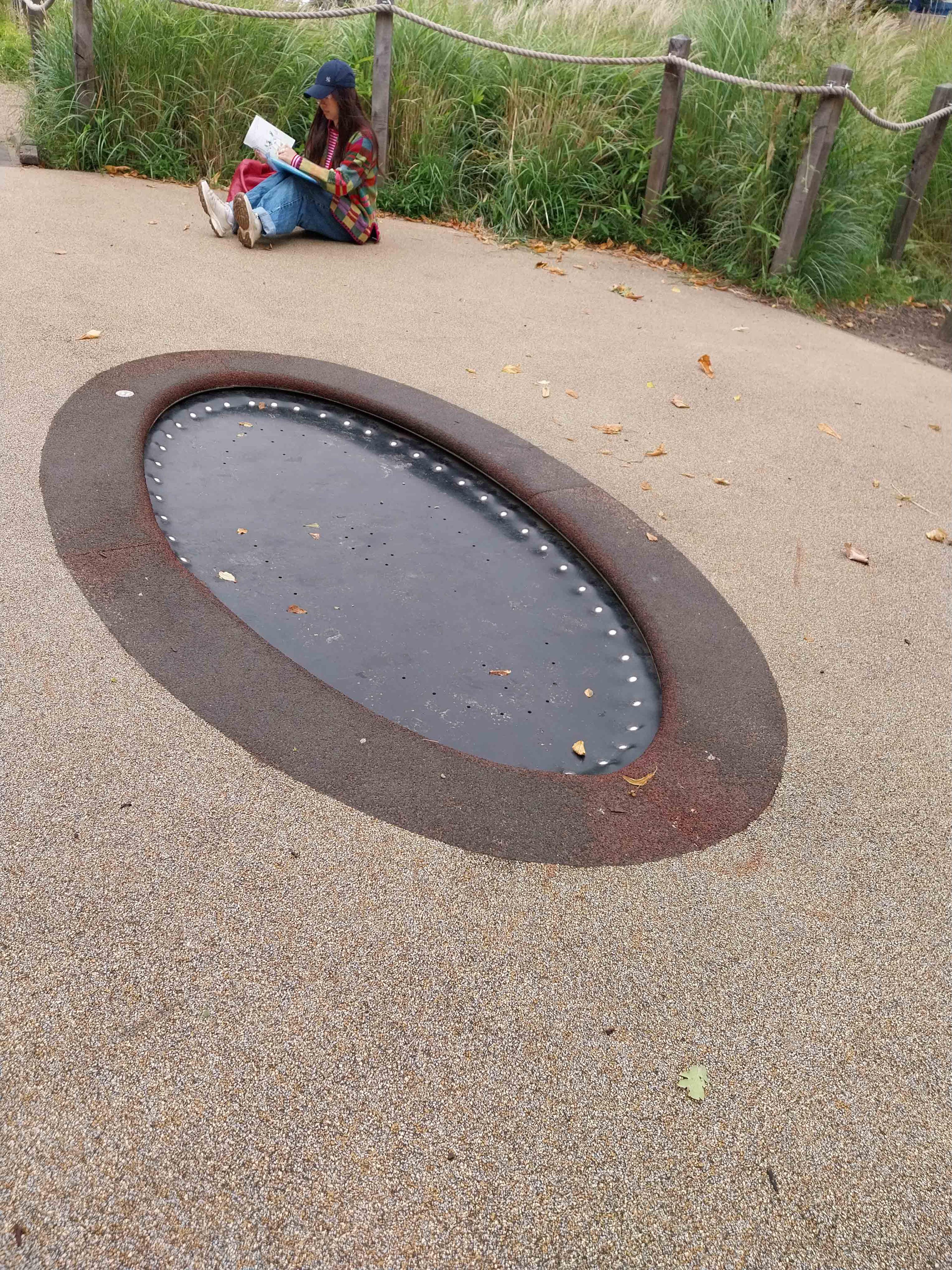 A person sitting on the ground illustrating, in the ground in front of them is a large circular trampoline.