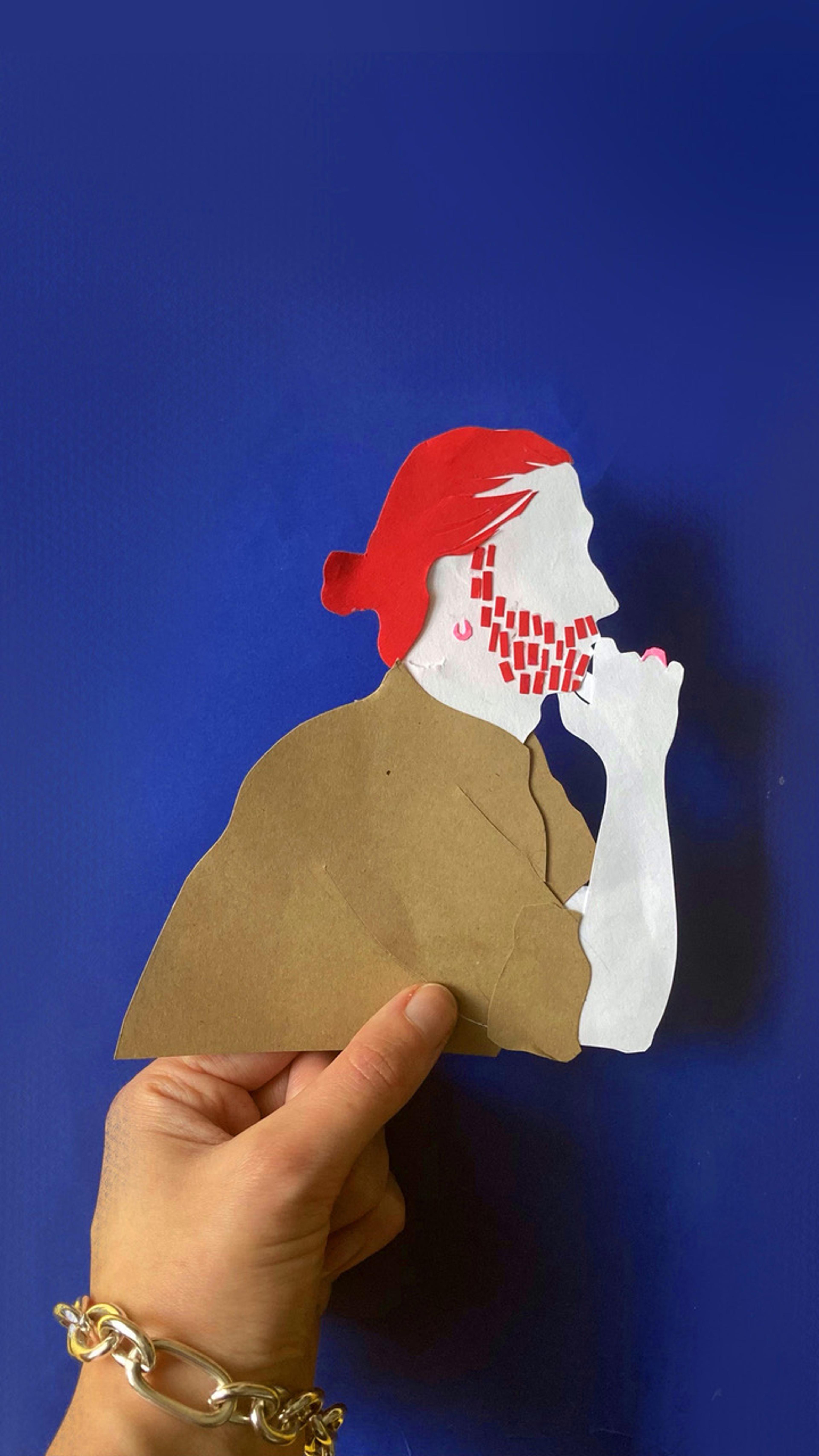 Photograph of a hand holding a paper cut out illustration of a person from the side with their hands under their chin.