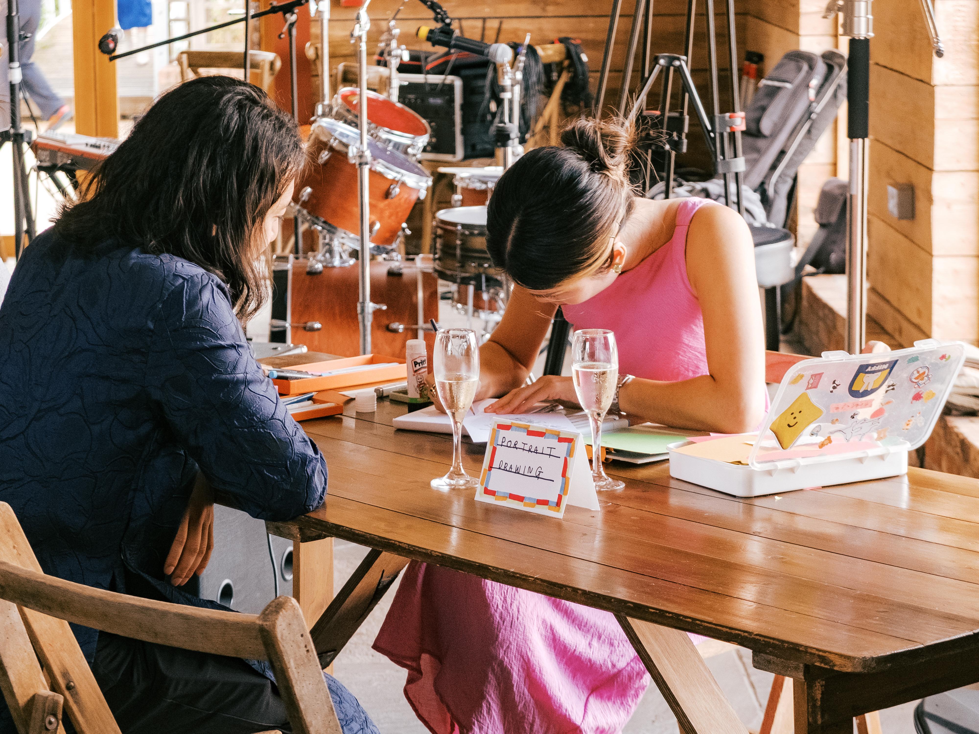 Two individuals sit at a wooden table with art supplies, champagne flutes, and a small box of stationery, with a drum set and filming equipment in the blurred background.
