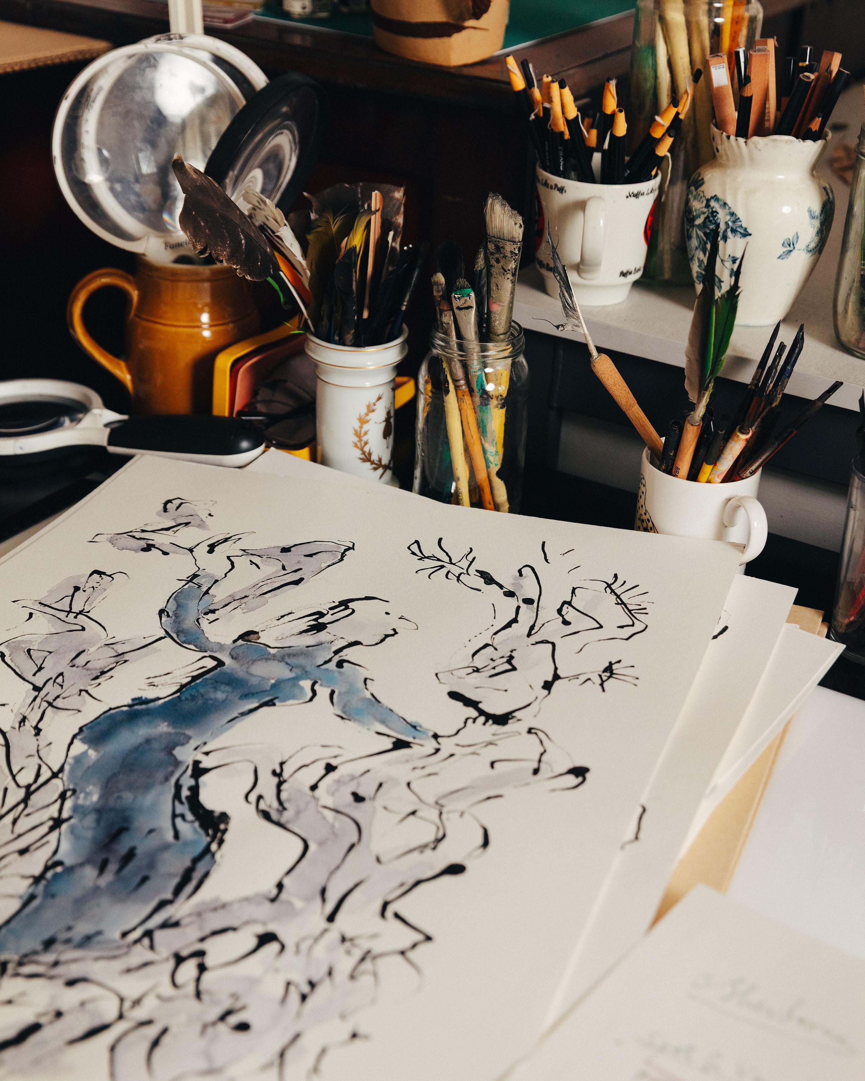 Pots of pencils and pens surround an ink and watercolour drawing of a woman dressed in blue surrounded by floaty foliage.