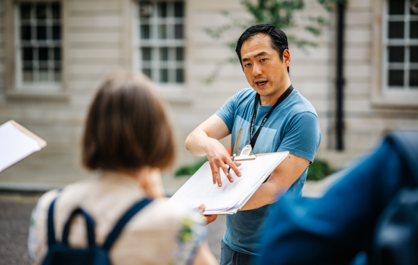 Man in a blue t-shirt points to a drawing on a clipboard while speaking to a person whose back is to the camera.