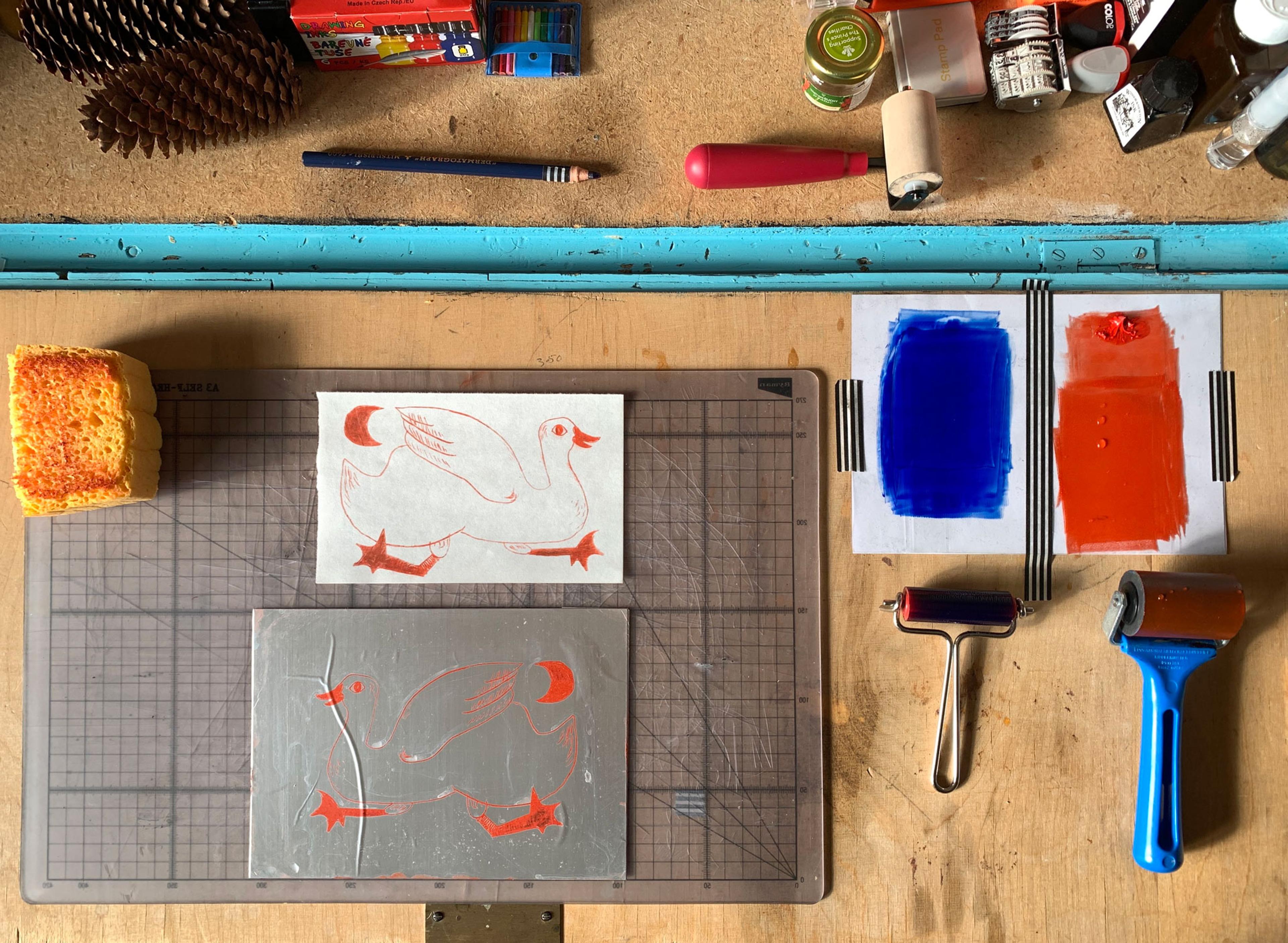 Ariel view of a desk where someone is printing with rollers, a sponge, a printing plate, art materials and paper.