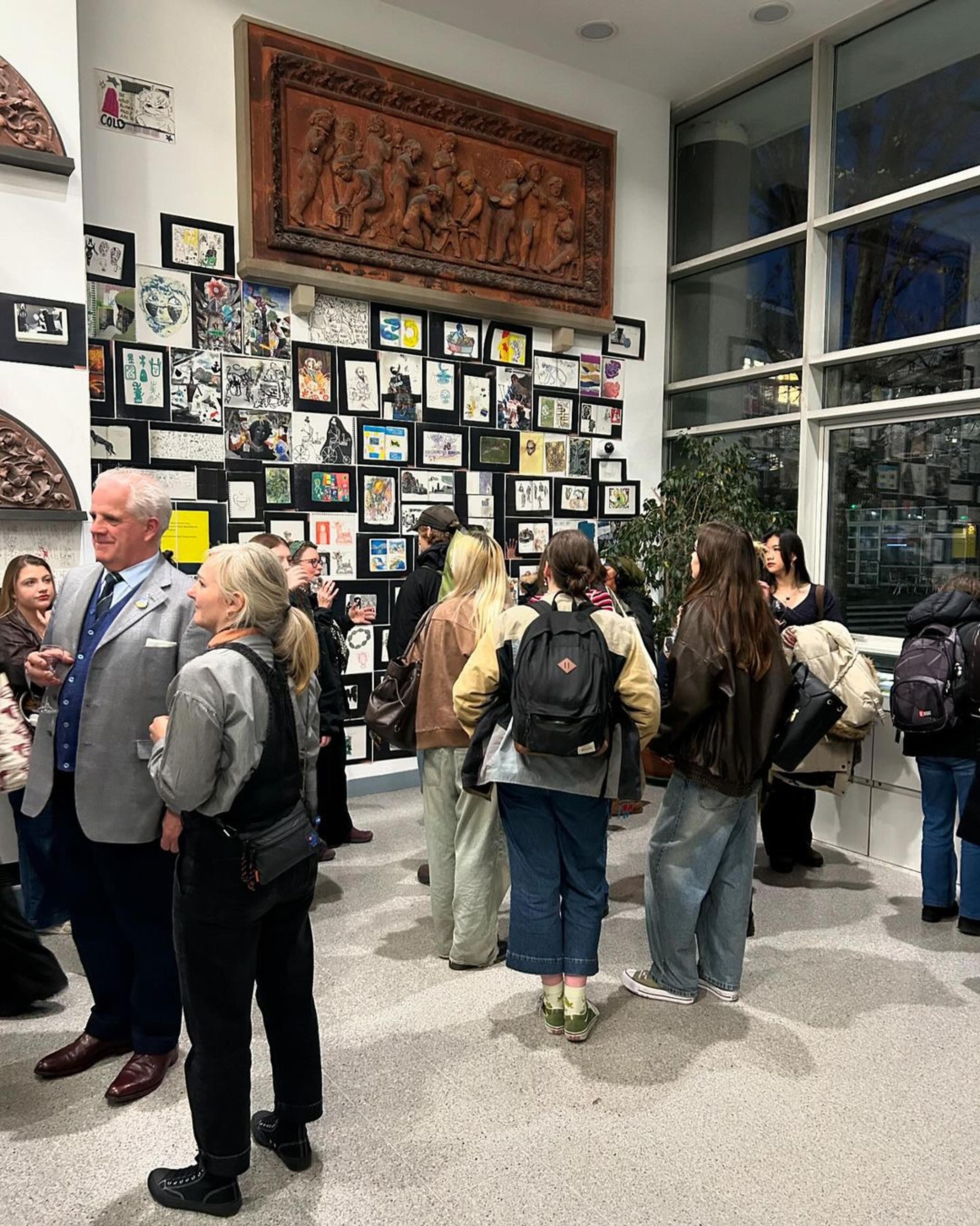 A crowd of people stood in a gallery space in front of an exhibition of illustrations displayed on a white wall 