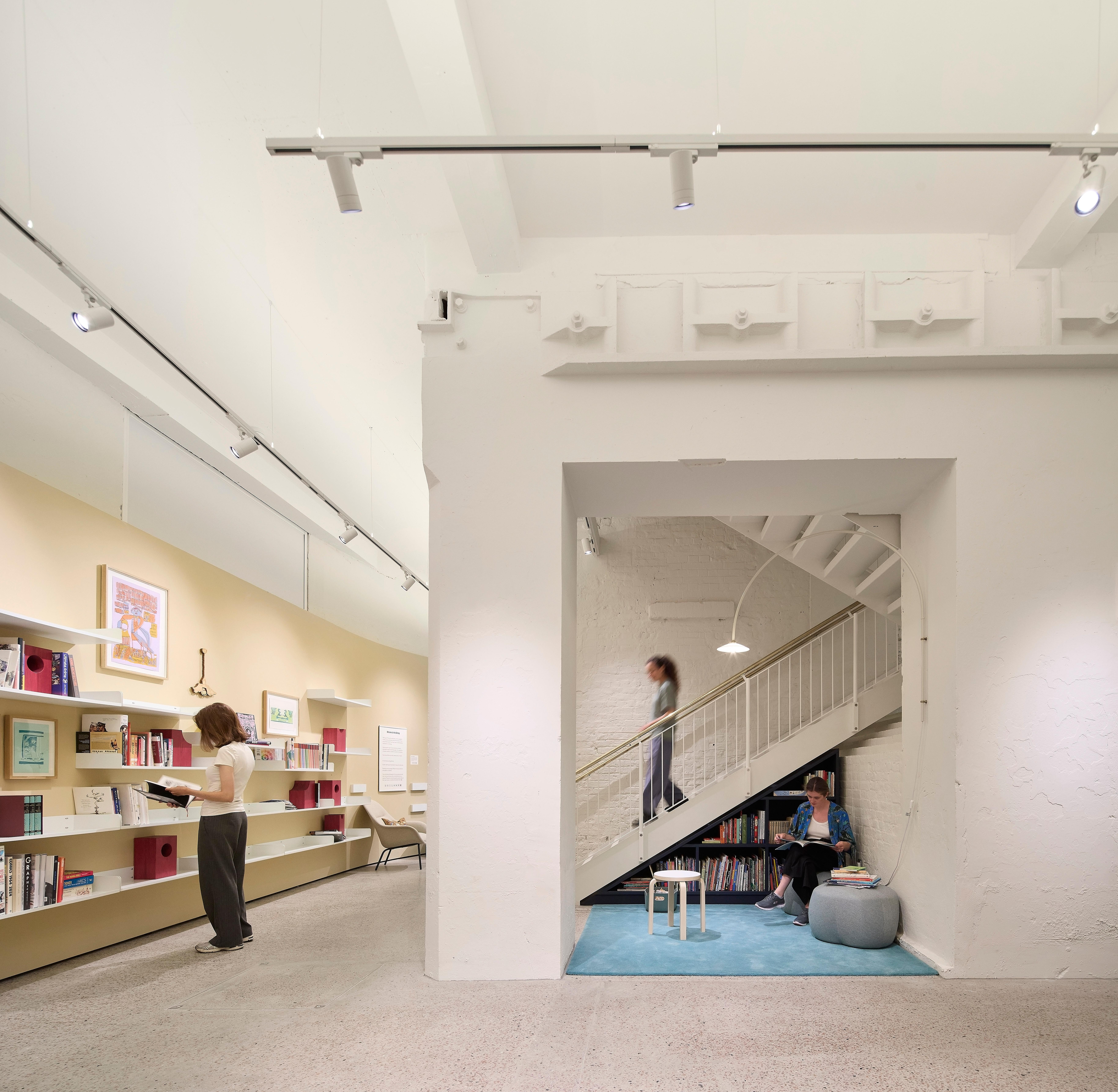 Woman walks down the stairs in a library as a woman reads books from the shelf.