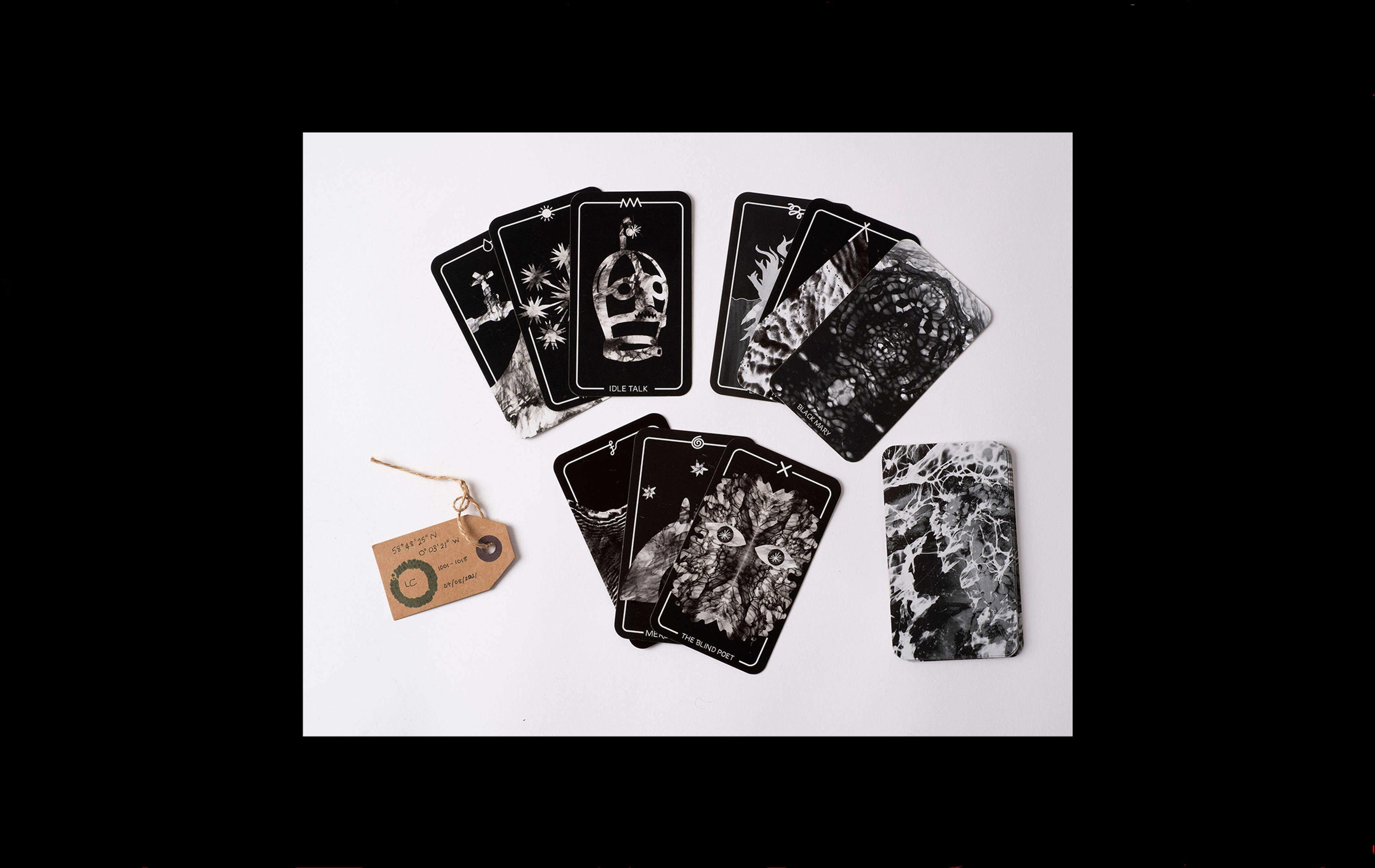 Photograph of back and white playing cards arranged in three hands. The top cards are 'idle talk' with an image of a scold's bridle, 'the blind poet' with two eyes and 'Black Mary' with an intricate pattern