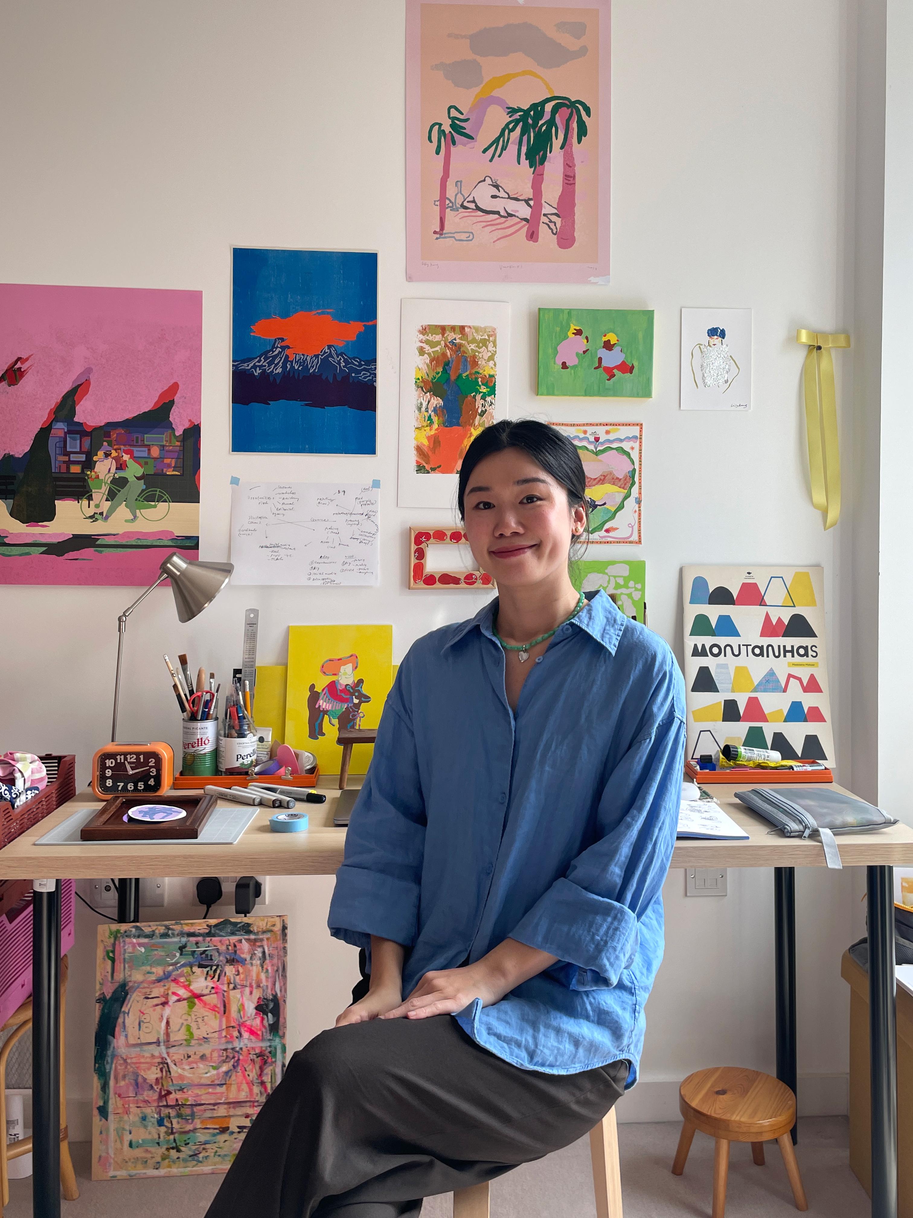 An artist sits at a desk in a studio, surrounded by colourful artwork on the walls and art materials.