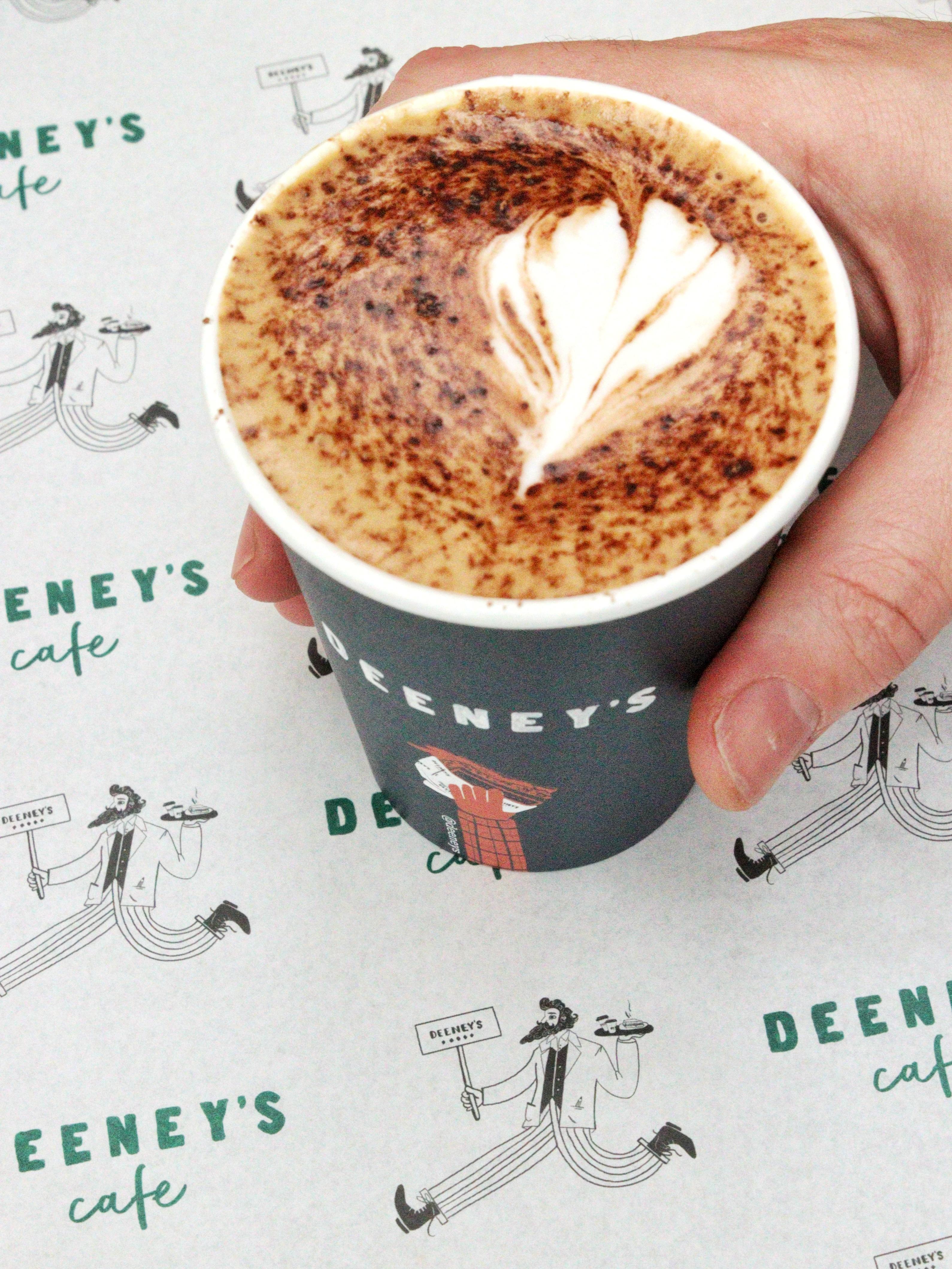 A frothy coffee with latte art and chocolate powder on top in a takeaway cup labelled Deeney's.