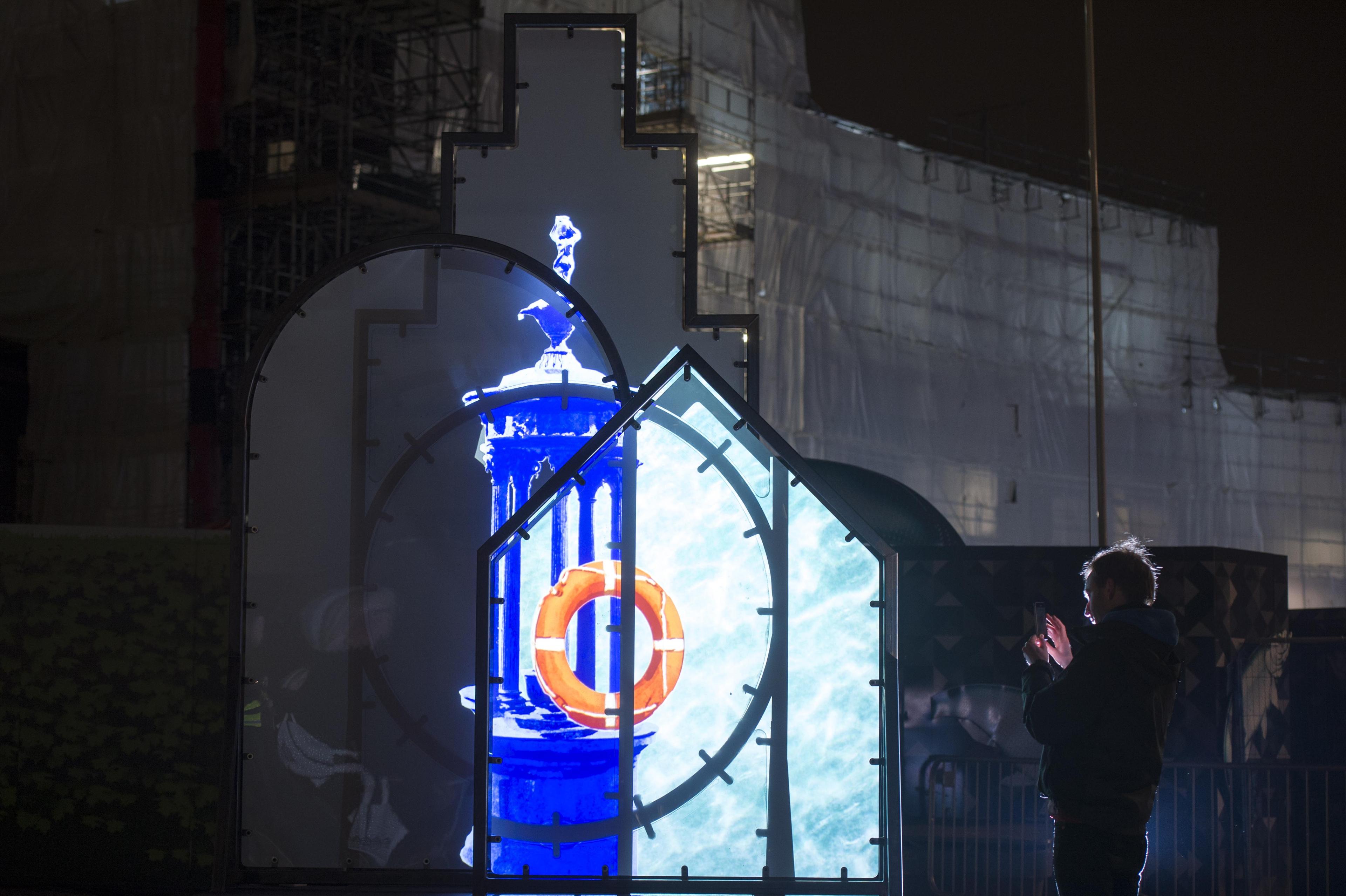 Art installation. Shapes installed outside with illustrations projected onto the shapes. A person is next to them is taking a photo of the installation.