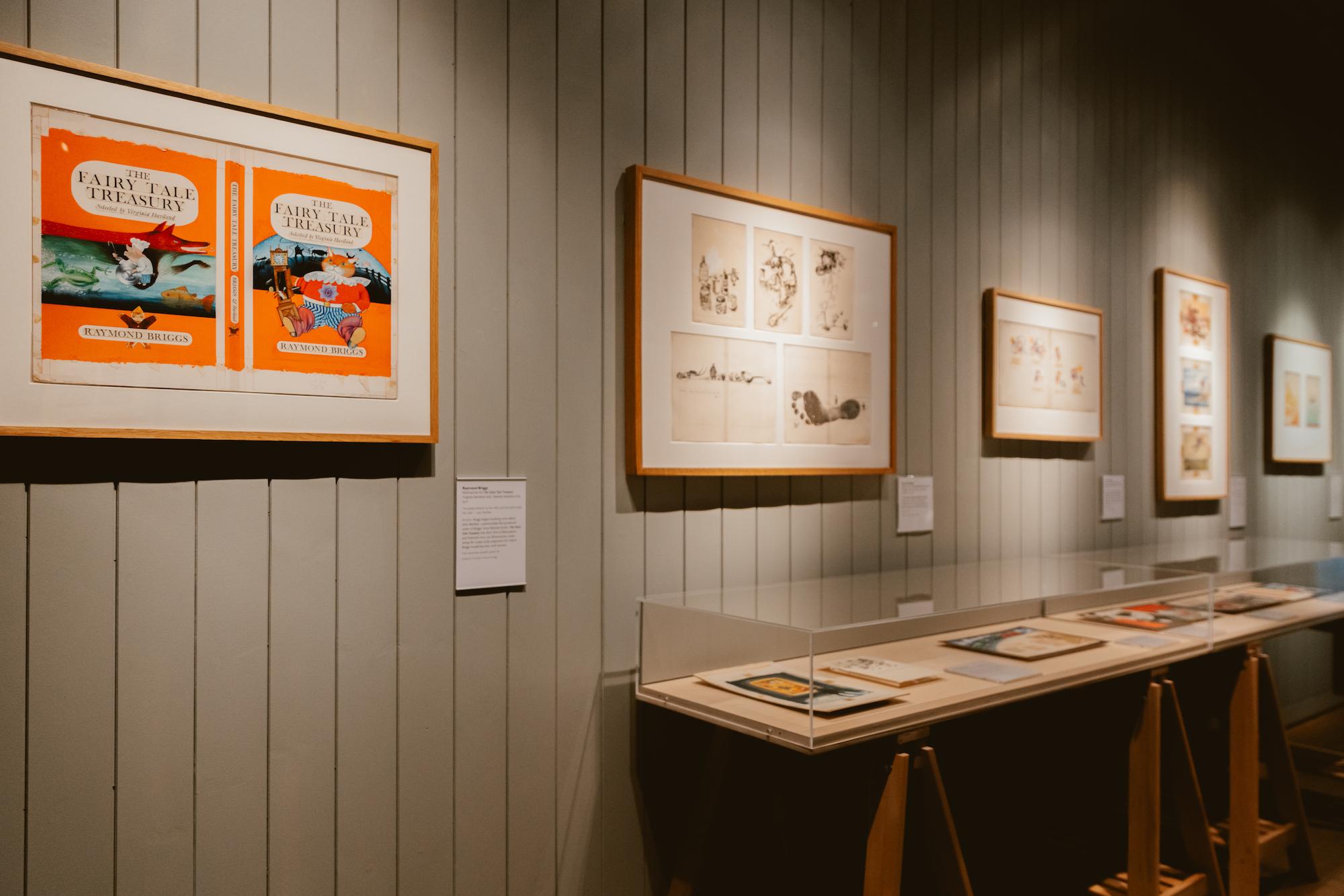 A photograph of a gallery with framed illustrations hanging on the wall and glass display cases against the wall
