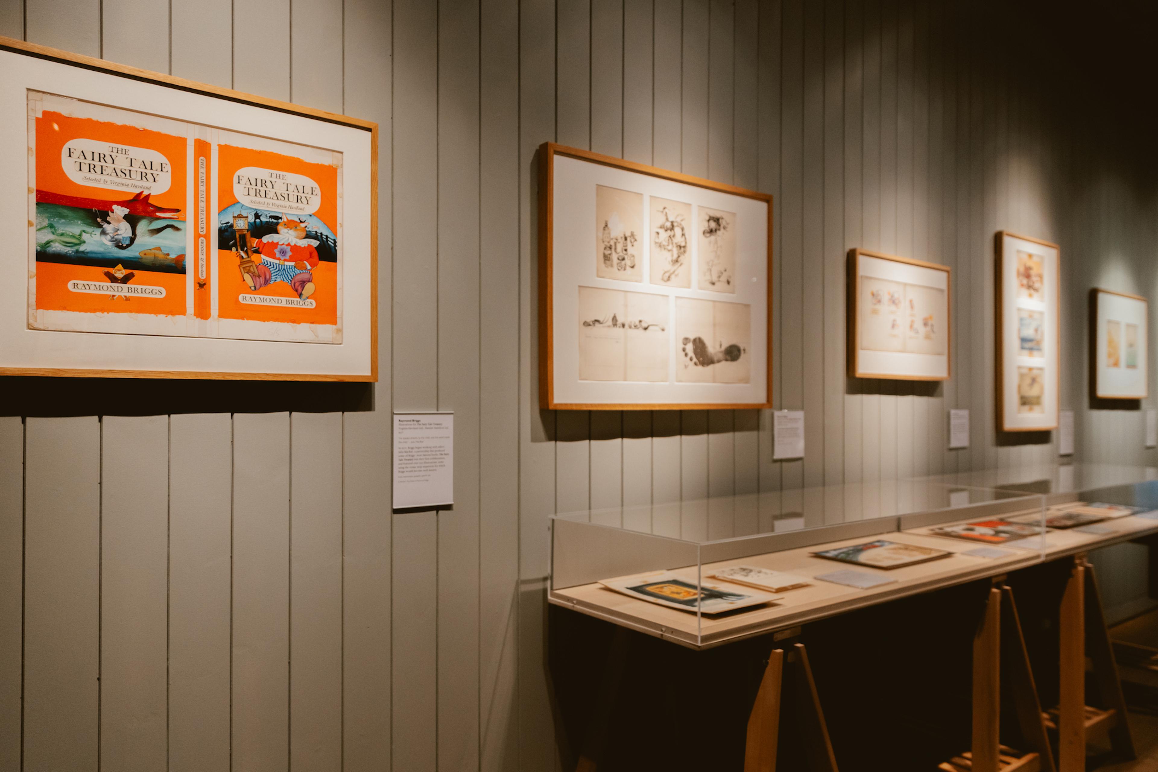 A photograph of a gallery with framed illustrations hanging on the wall and glass display cases against the wall