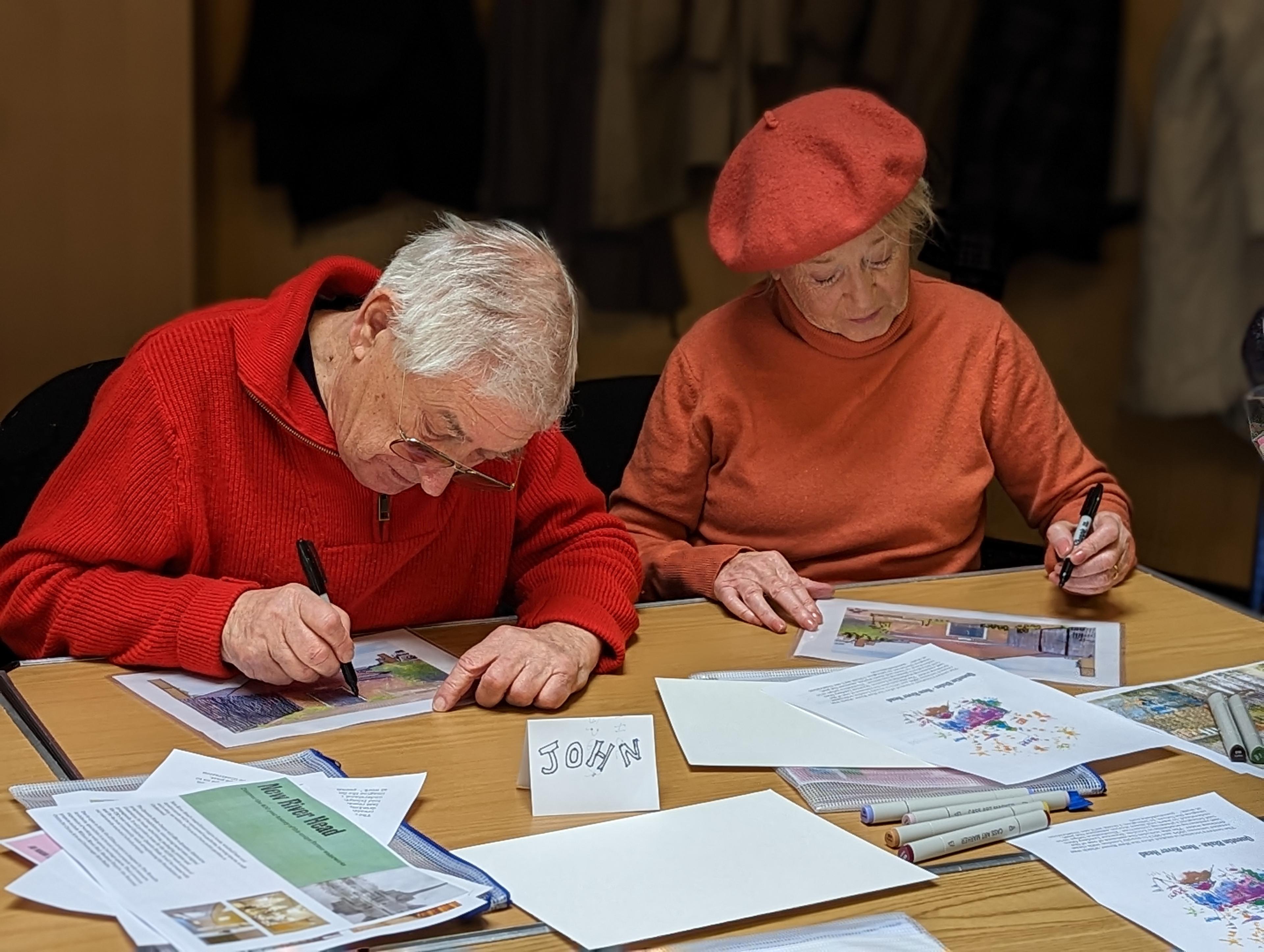 Two older adults sat at a laminated table, drawing with markers.