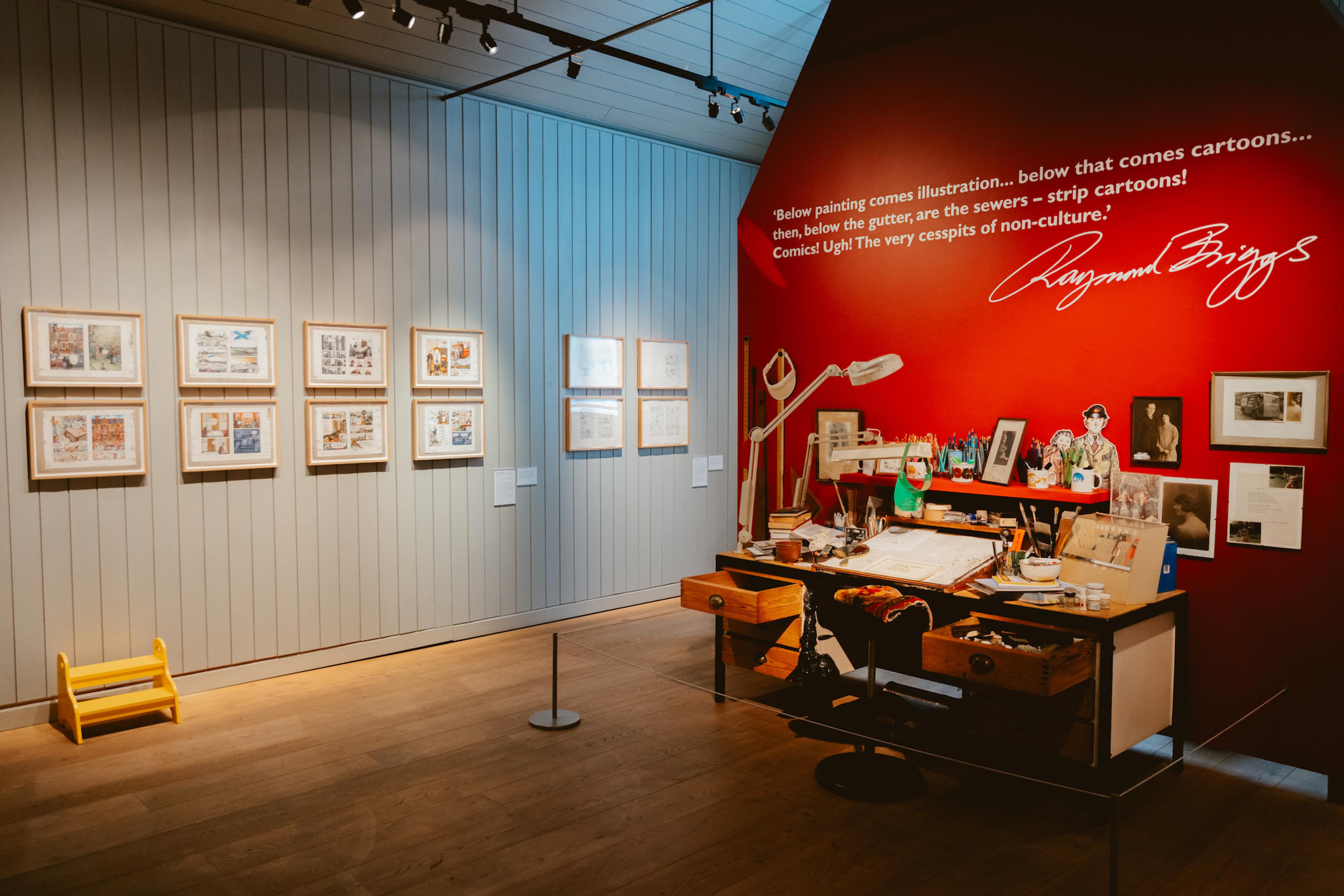 A photograph of a gallery with framed illustrations on the walls and an exhibit of the illustrator's desk and chair against one wall 