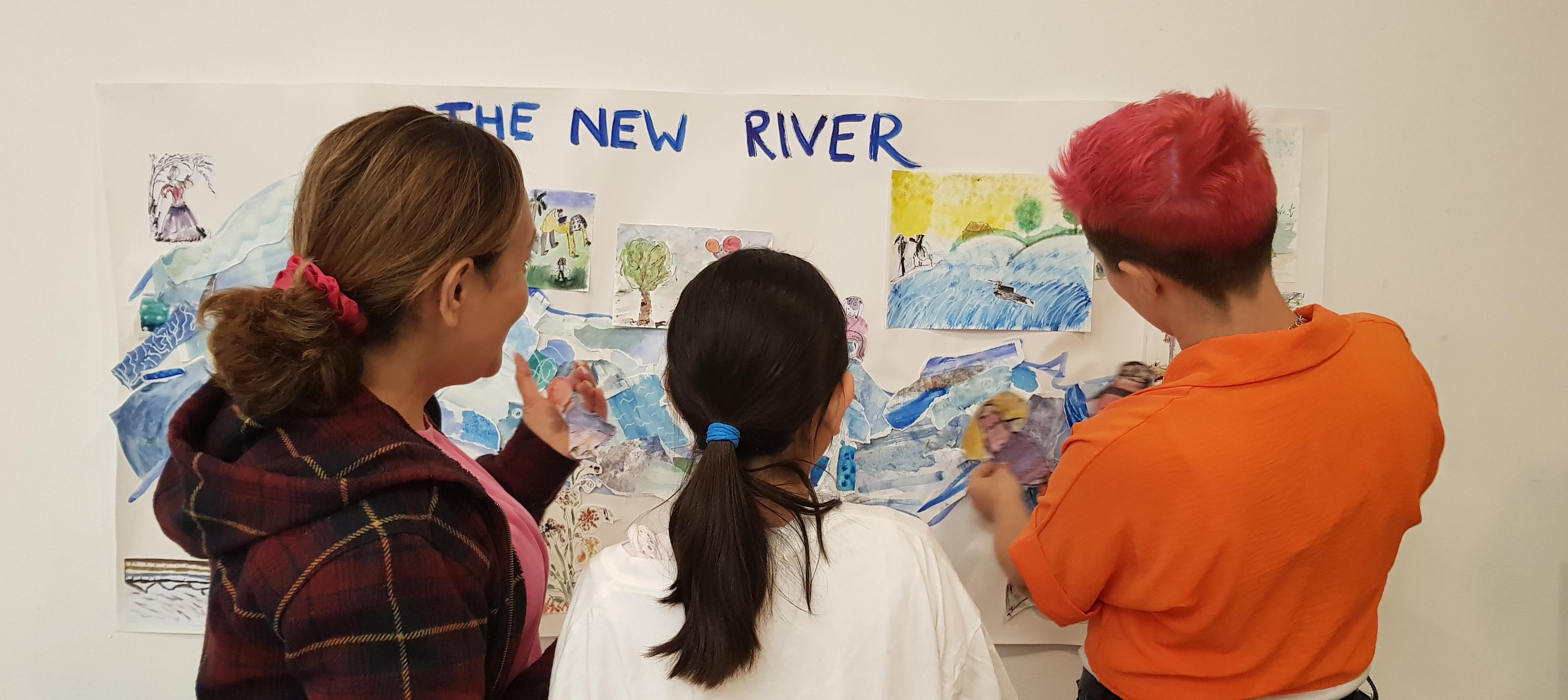 Photograph of the back of three people standing in front of a wall, displaying paintings on the wall. The words New River Head is painted onto a piece of paper on the wall.