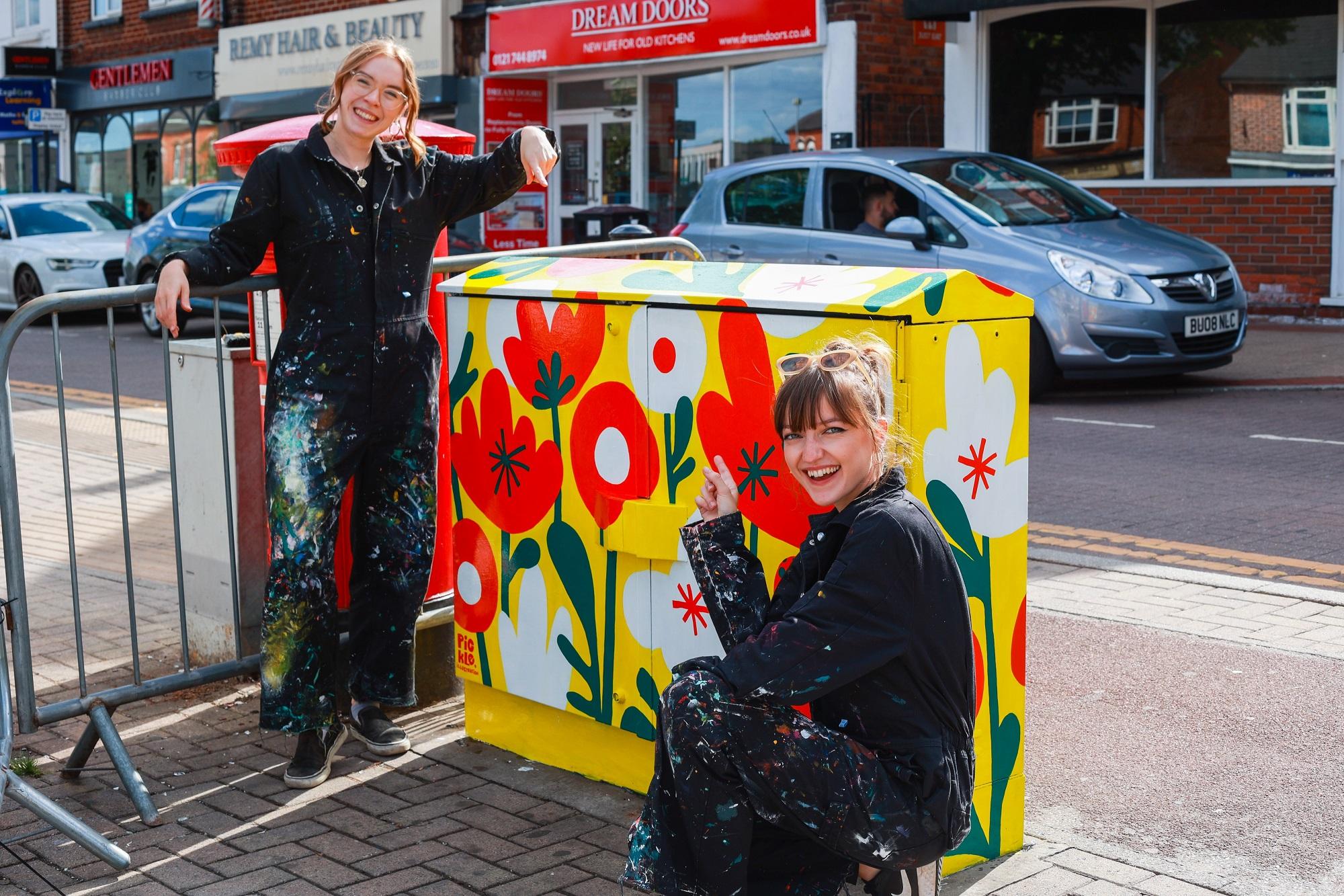 Two people in overalls, pointing at a utility box on the street that has been painted in bright yellow with orange and white flowers.