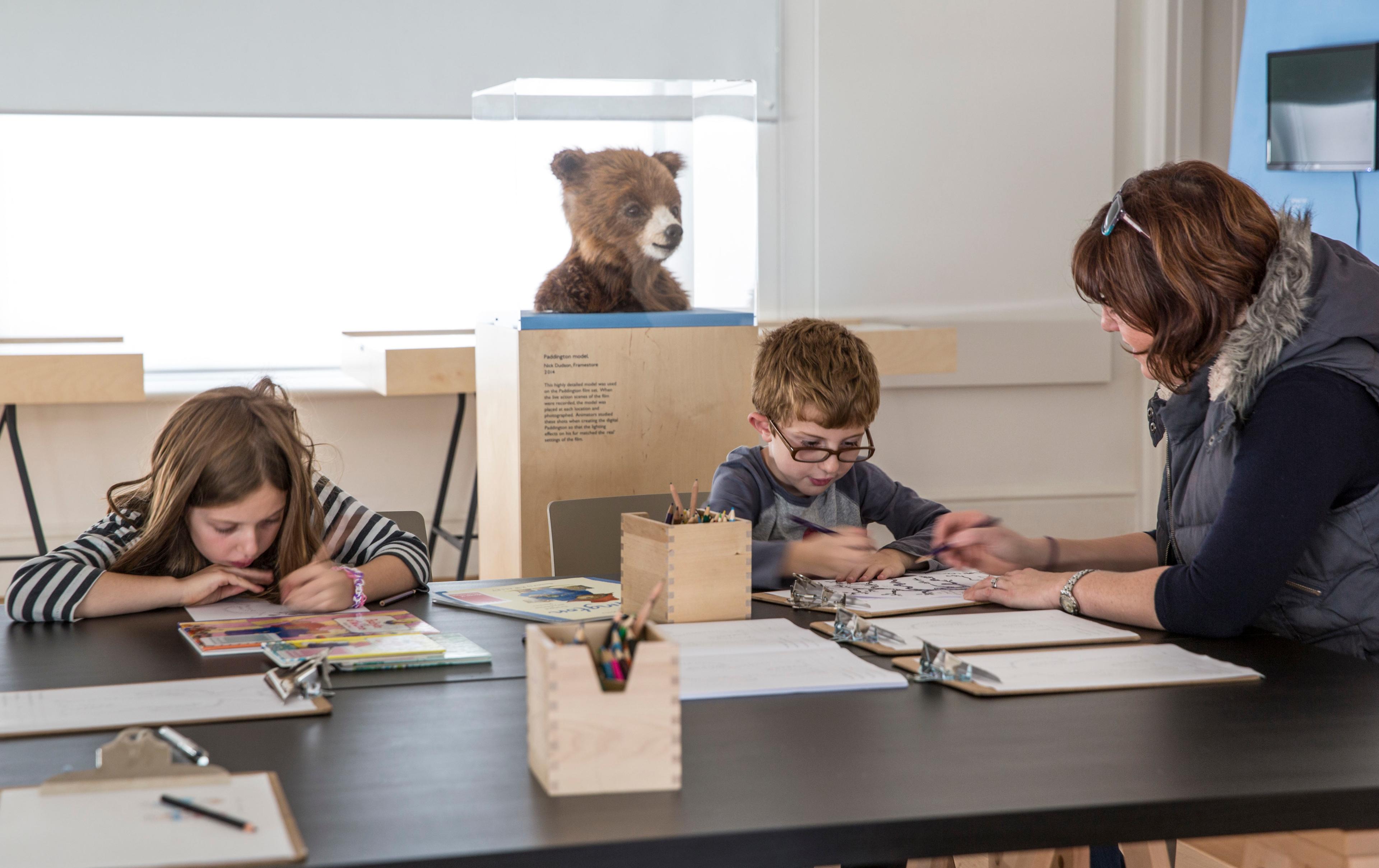 An adult and two children illustrating at a table, in the background there is a furry bear head is in a glass display case.