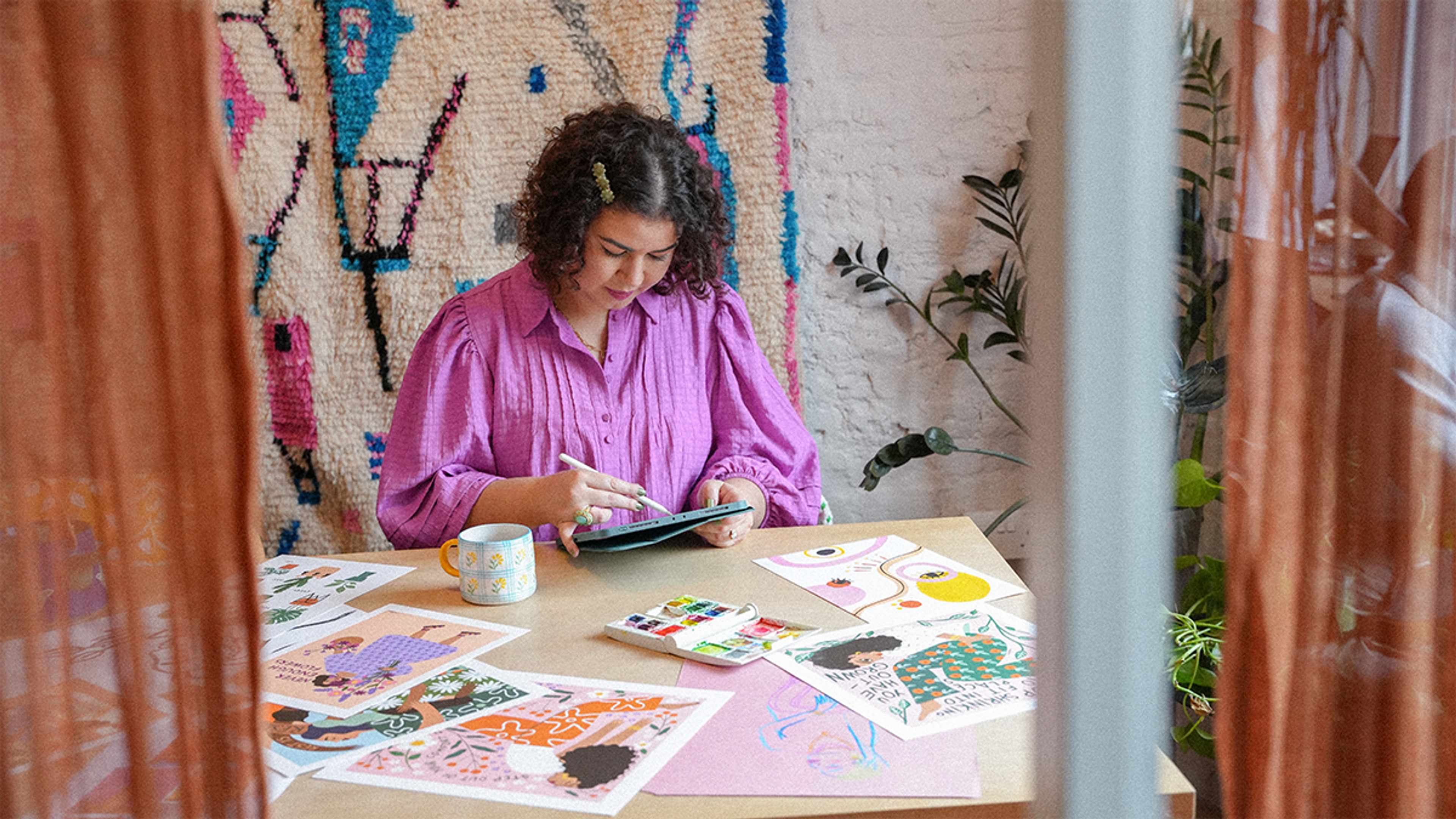 A photograph of a young woman sat at a desk drawing on a piece of paper. The desk is covered in prints of colourful illustrations.