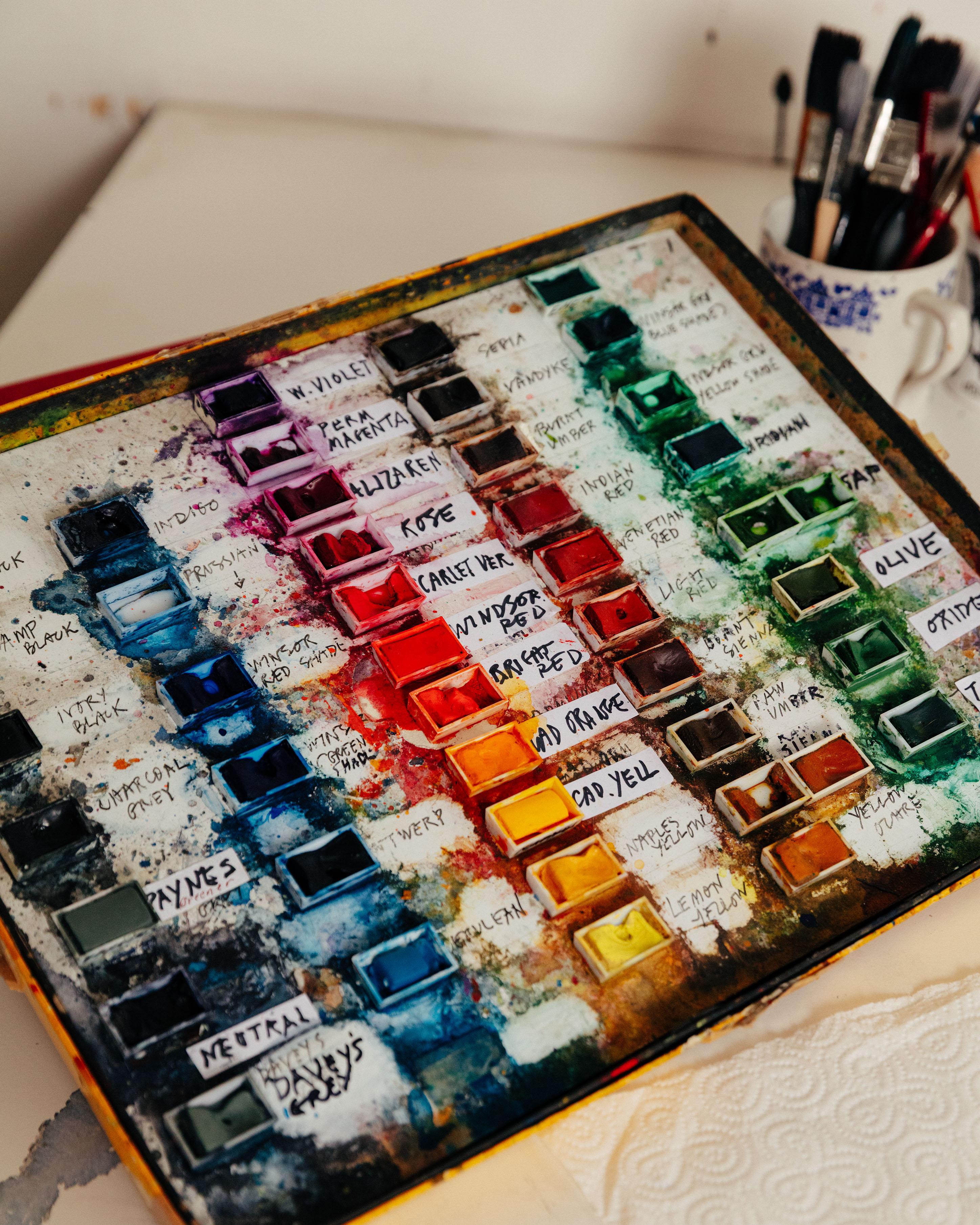 A colourful watercolour paint palette with a pot of pens and brushes in the background.