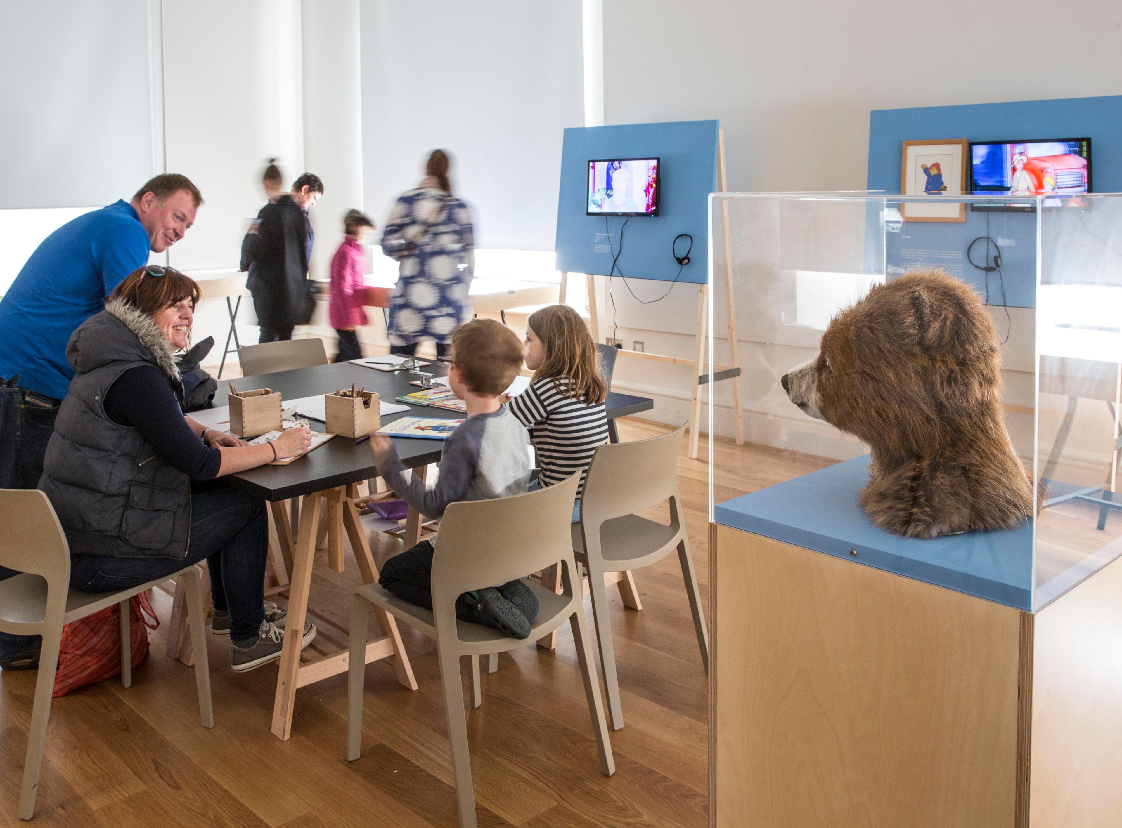 Adults and children in an exhibition space with tables, chairs and displays including a furry bear head is in a glass display case. 