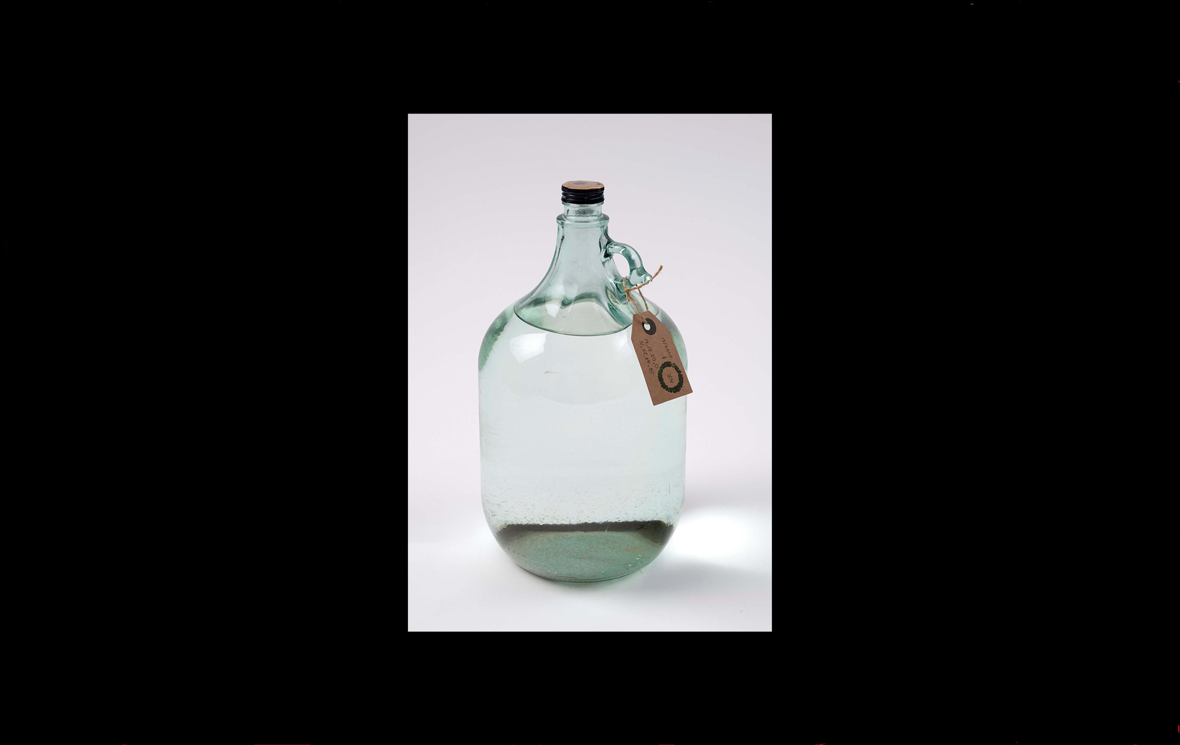 Photograph of a large glass jug full of water with a museum label around the neck