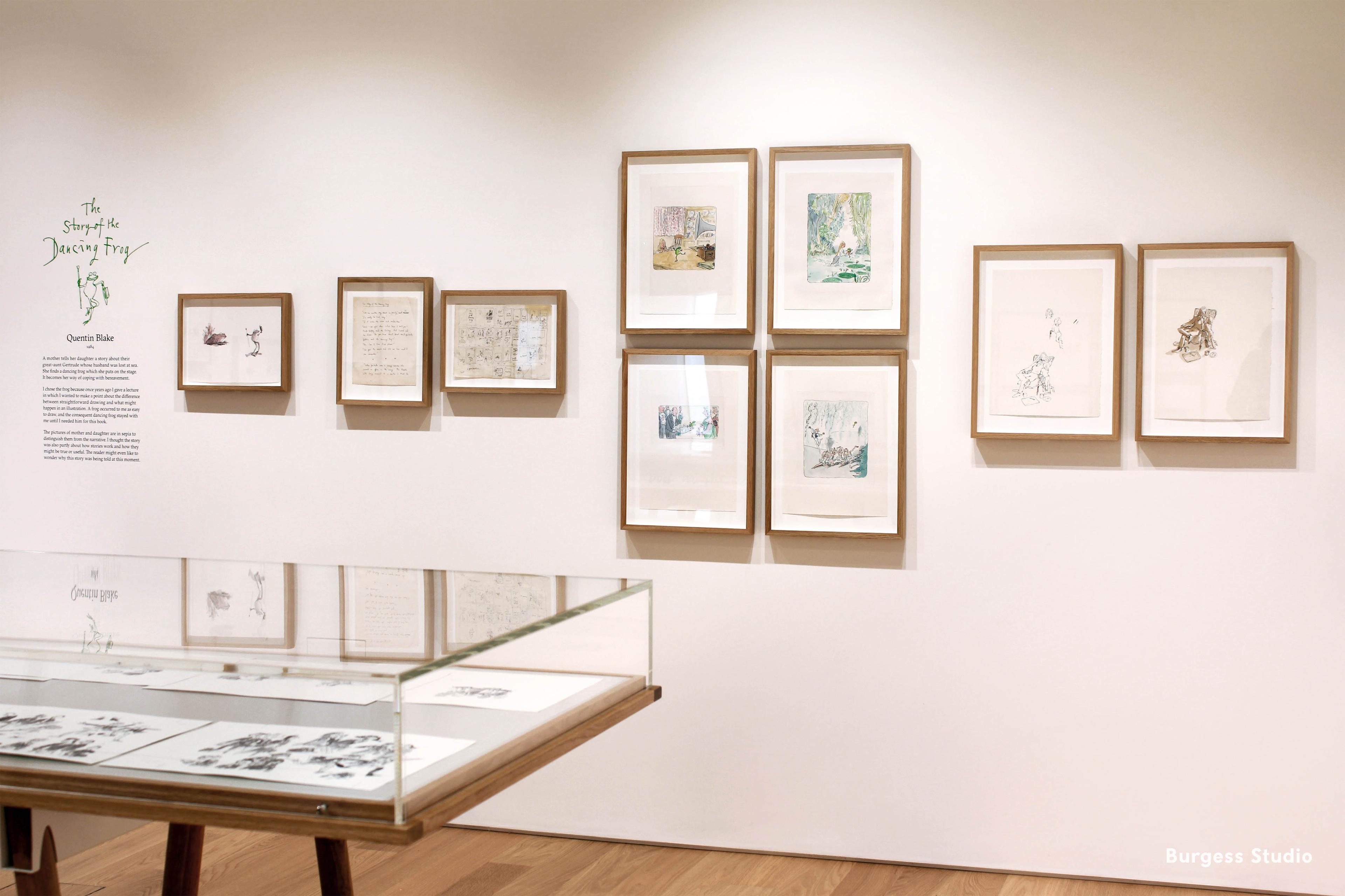 Inside an exhibition space with framed illustrations on the wall and displayed in a glass cabinet.