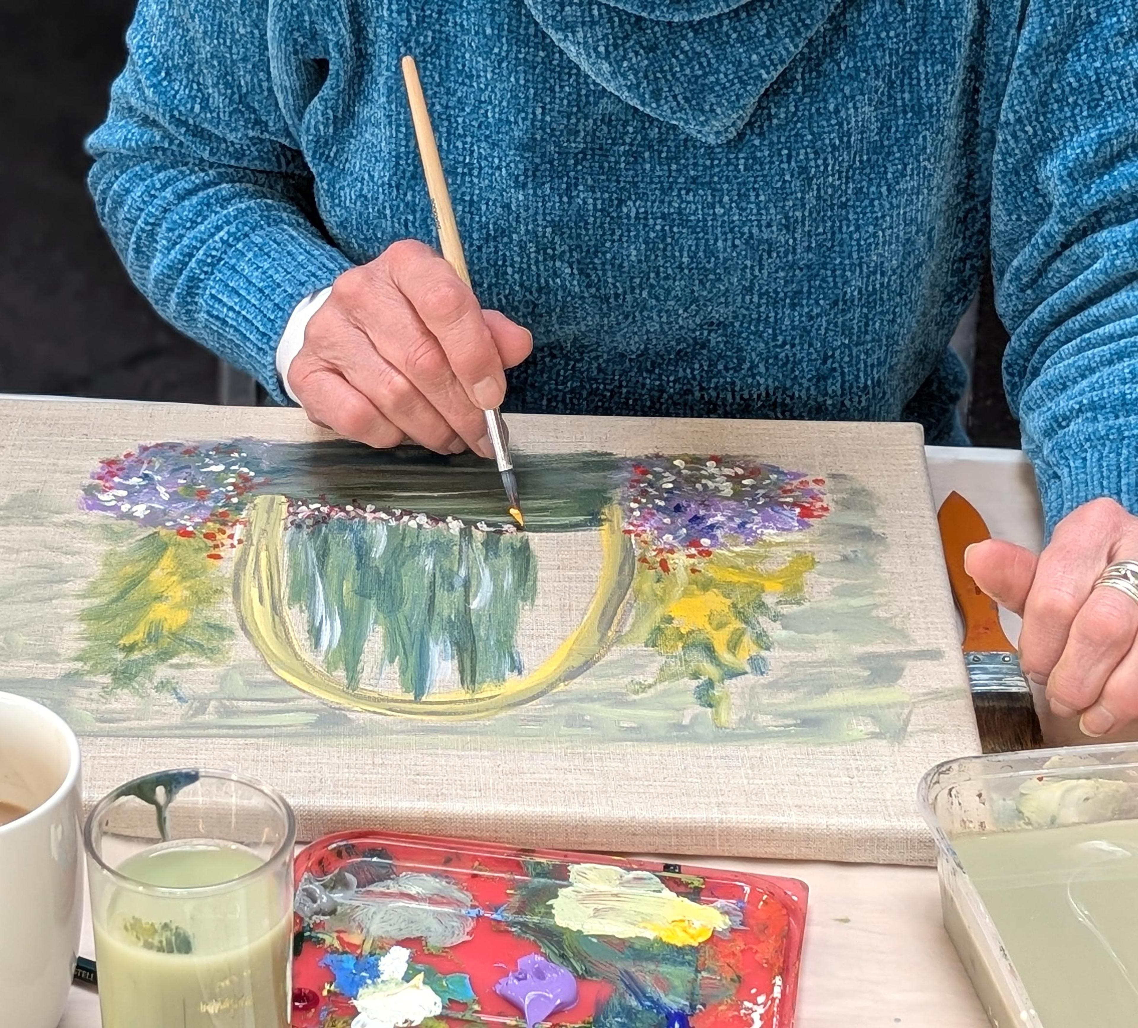 A photograph of a fabric canvas and paint palette on a table. An adult's hand is holding a paintbrush and painting a scene with foliage and flowers on the canvas. 