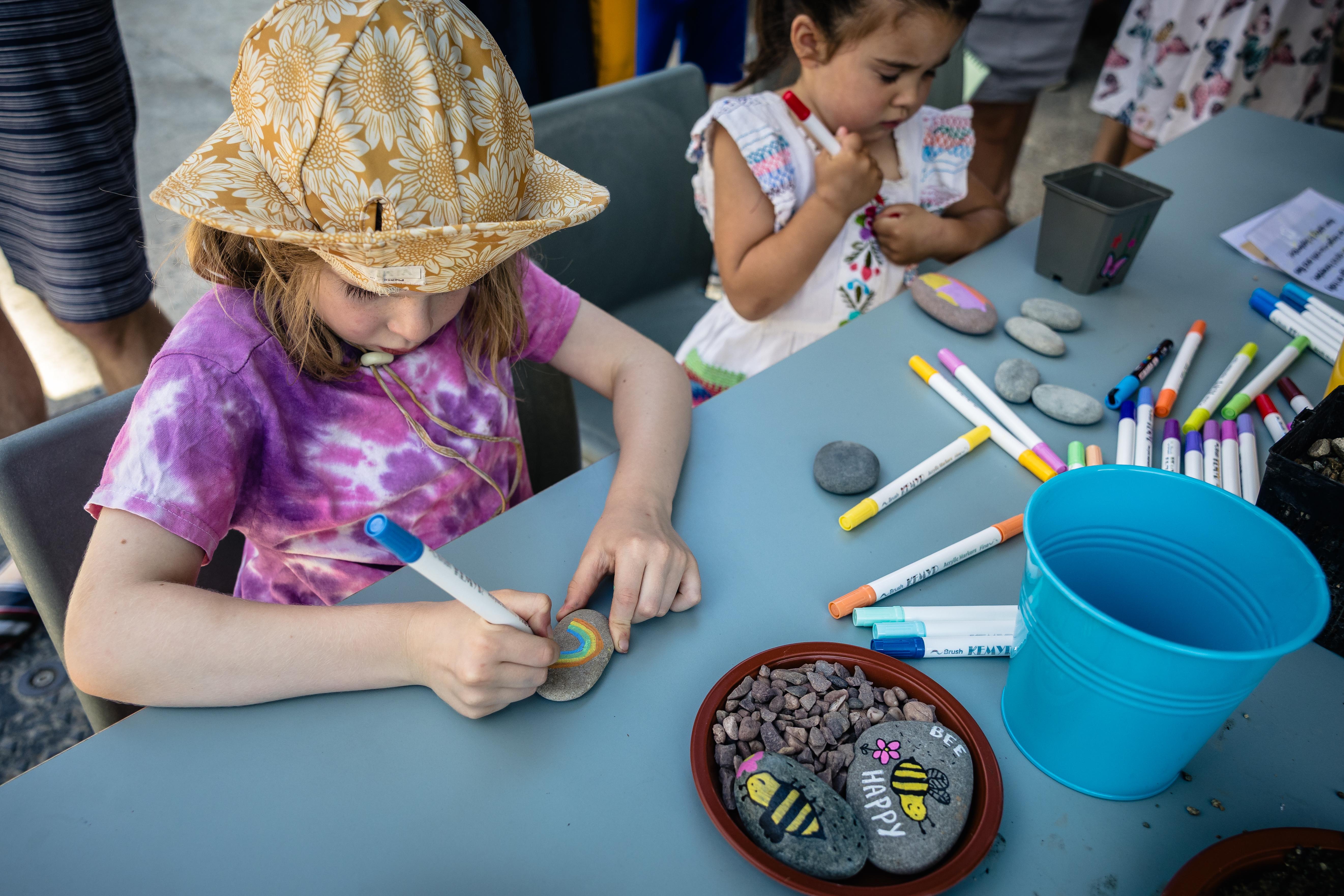 On a blue table, two children use colourful felt-tip pens to draw onto stones. Two stones on the table are illustrated with bees, one reading "Bee happy".